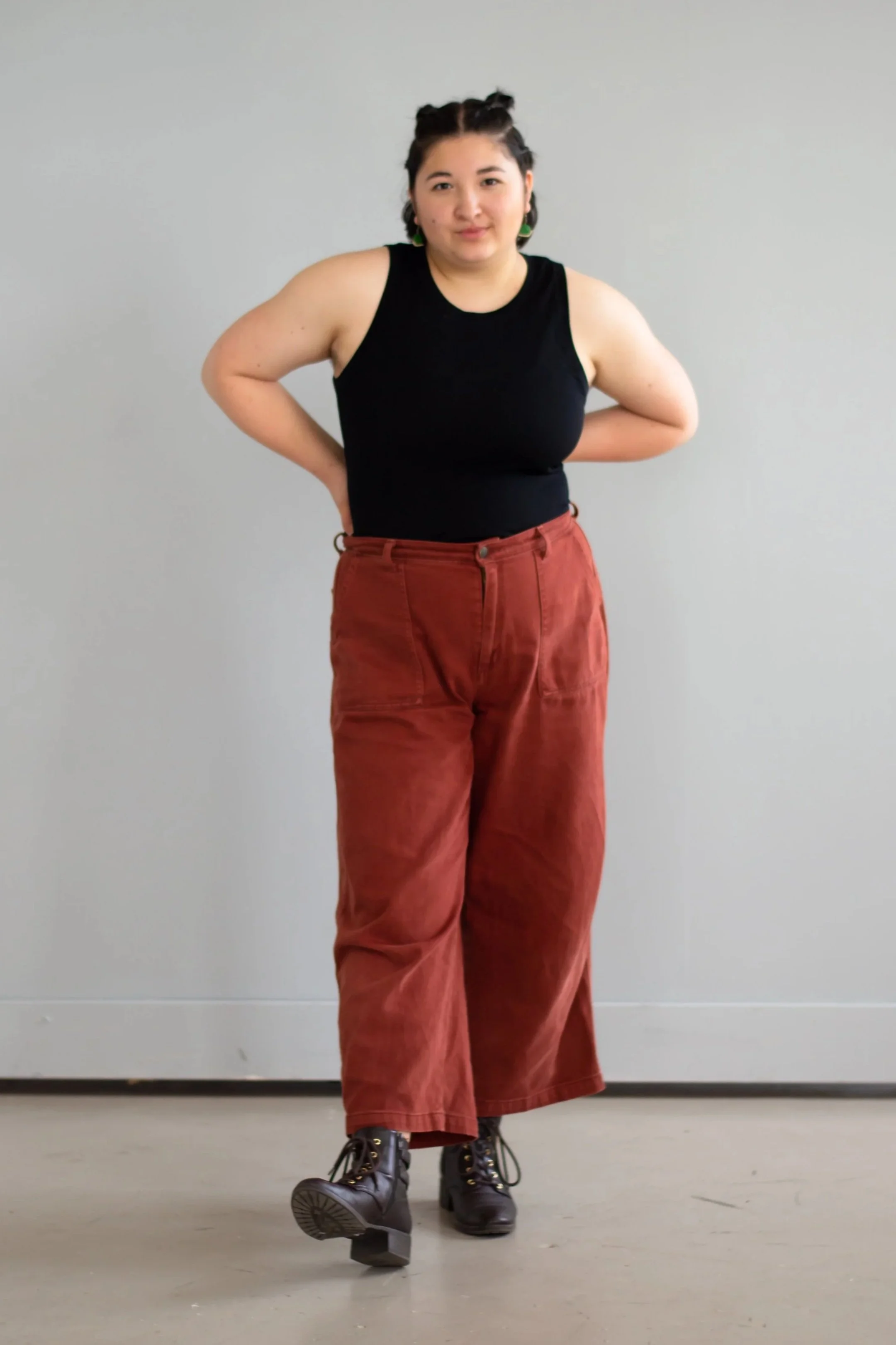 An Asian stage and theater director standing against a plain wall wearing a black tank top, red pants, combat boots, and colorful earrings, with arms behind their back.