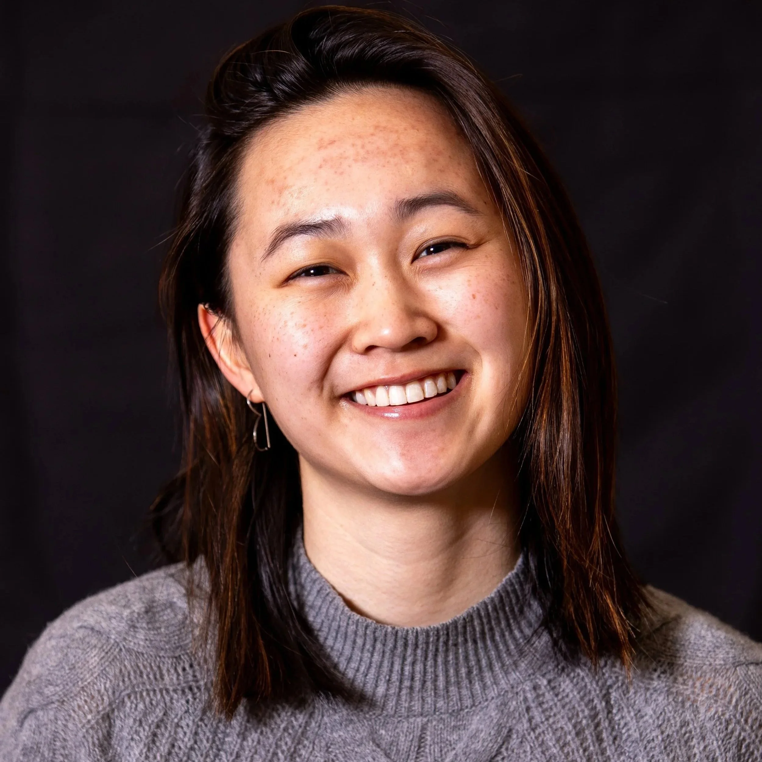 Close-up of asian theater and stage director with dark brown hair and wearing a gray turtleneck sweater, against a black background.