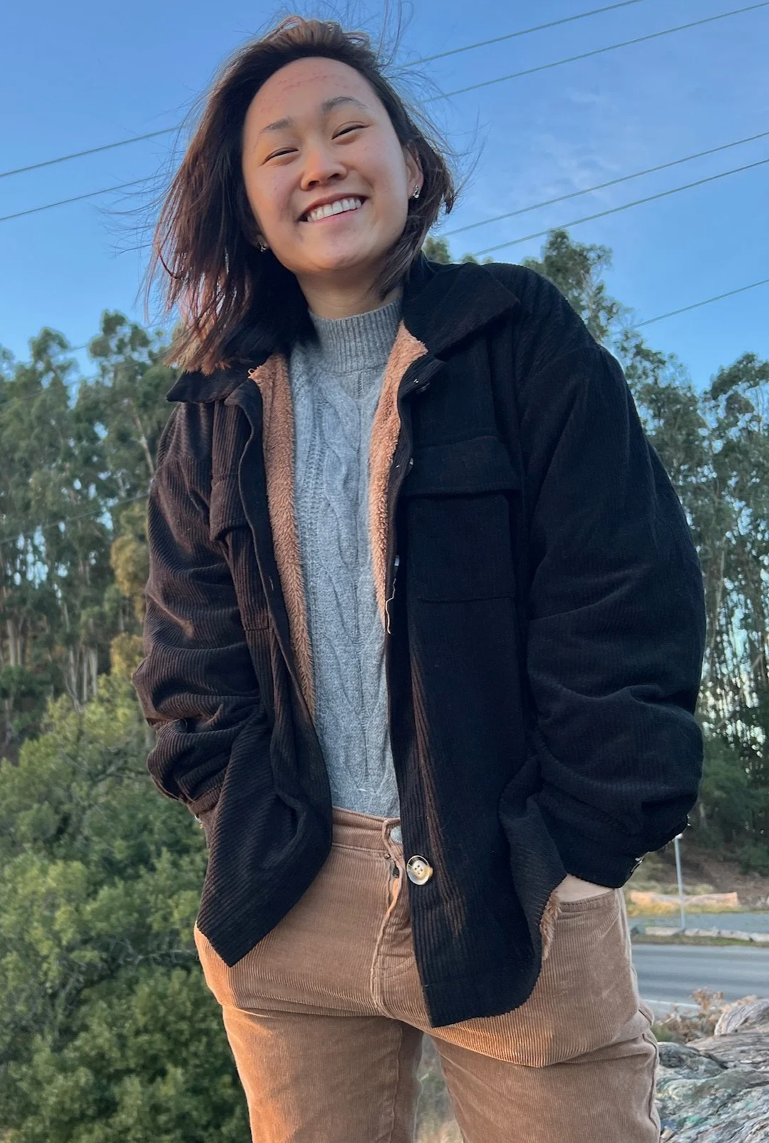 A smiling Asian stage and theater director with short hair wearing a black jacket over a gray sweater, standing outdoors with trees and power lines in the background.