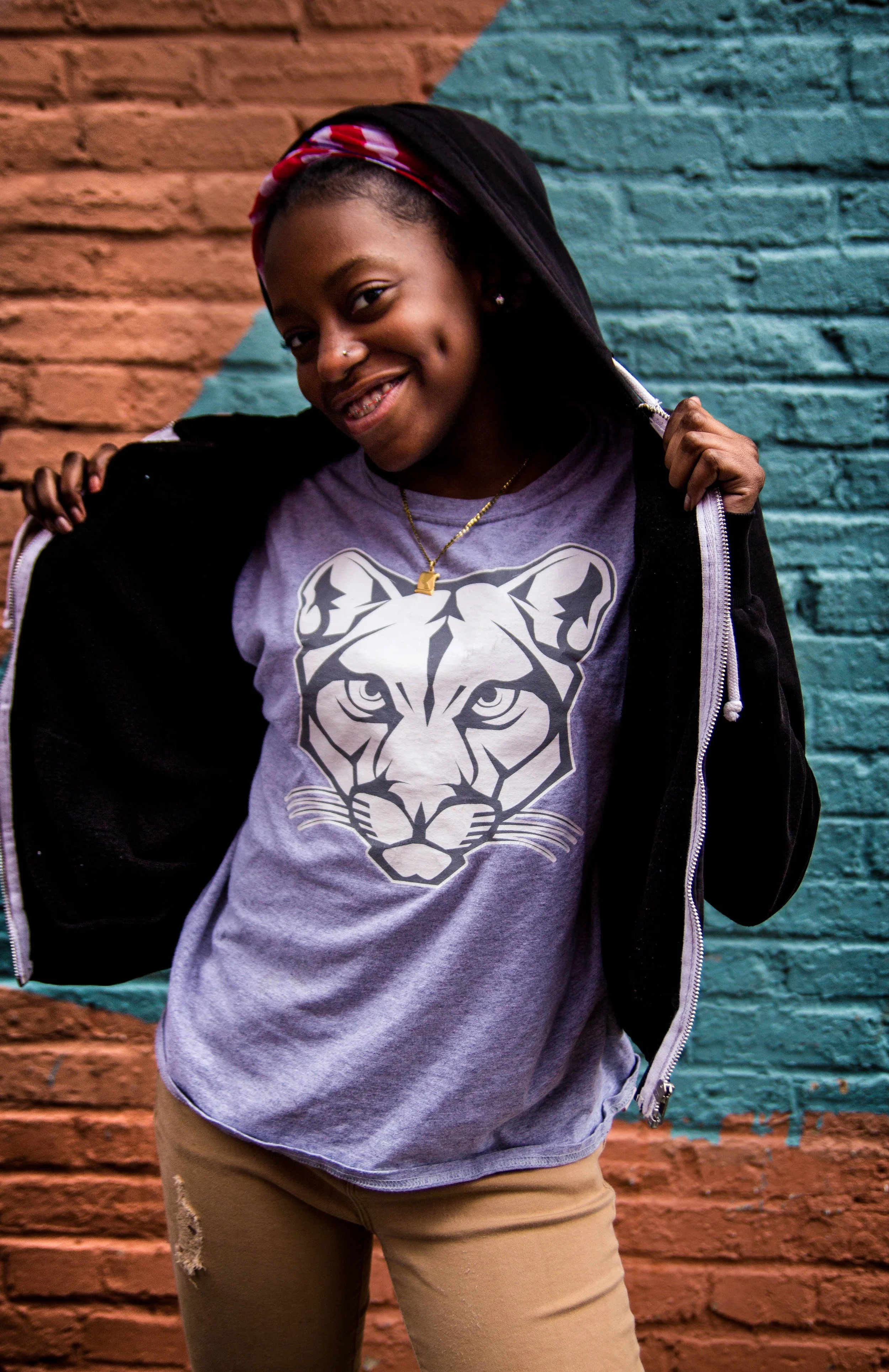 A young woman standing in front of a brick wall painted with blue and orange sections, wearing a gray T-shirt with a tiger face graphic, khaki pants with ripped knees, a black jacket, and a red patterned headband, smiling and holding the hood of her jacket.