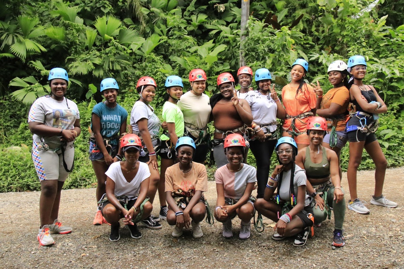 A group of women and girls wearing helmets and harnesses, posing outdoors in a lush green jungle setting after a rock climbing or ziplining activity.