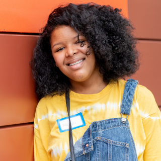Young girl with curly hair smiling, wearing a yellow tie-dye shirt, denim overalls, and a name tag, standing in front of an orange panel background.