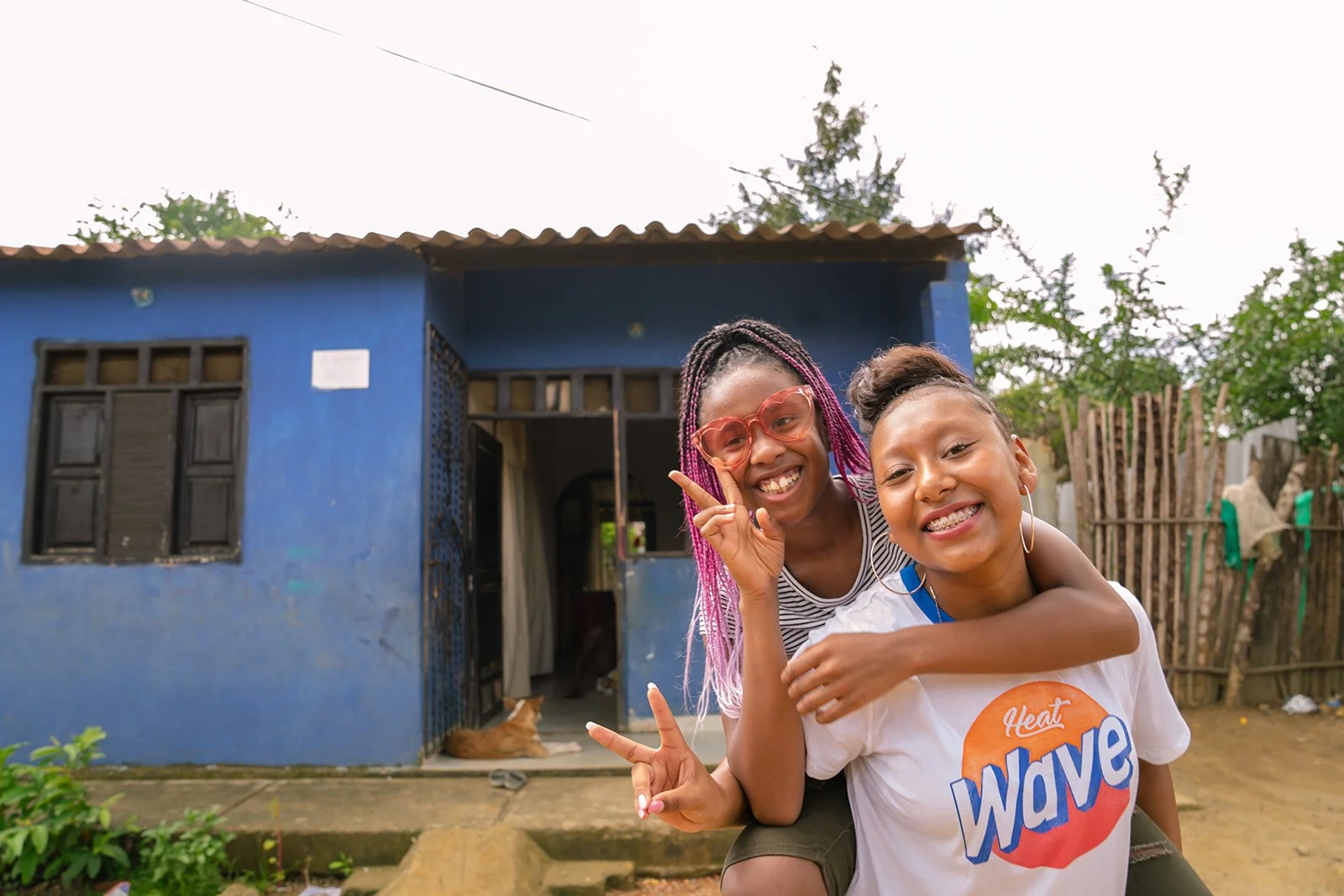 Two smiling girls with their arms around each other posing in front of a blue house with an open door, with a dog resting inside, in a rural setting with trees and plants.