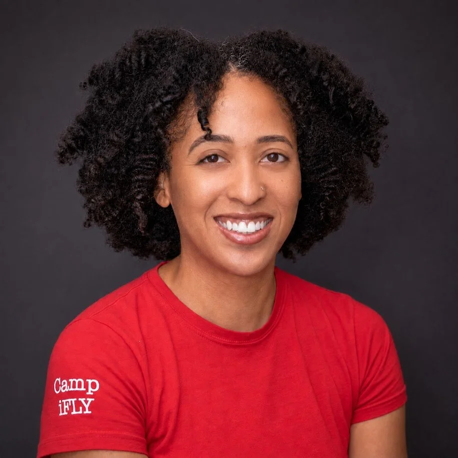 A young woman with curly hair wearing a red t-shirt with 'Camp iFLY' written on the sleeve, smiling against a dark background.