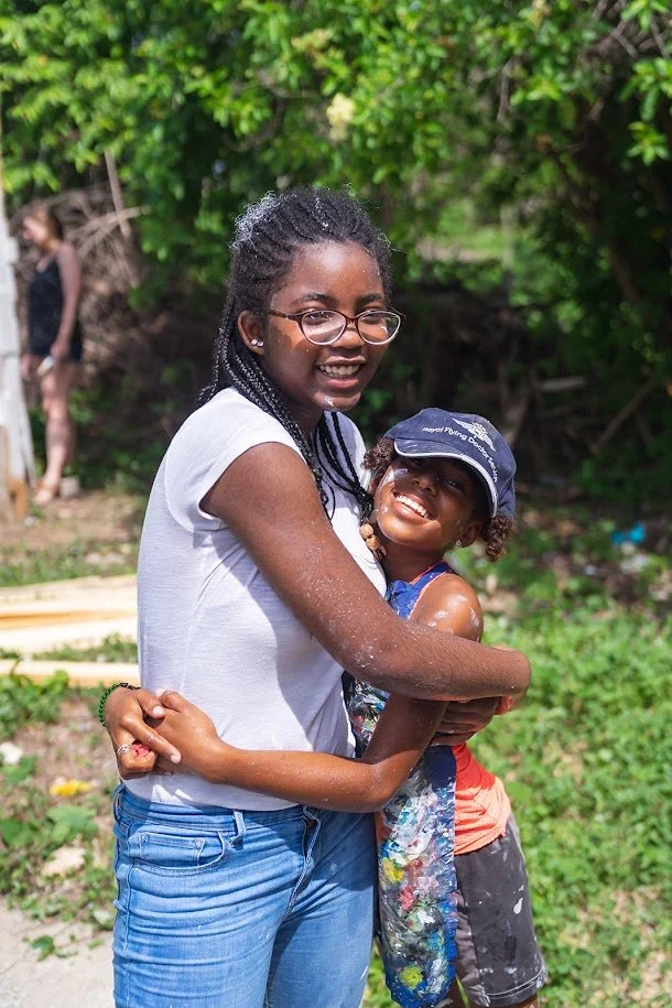 A woman and a young girl hugging outdoors with green foliage in the background, both smiling.