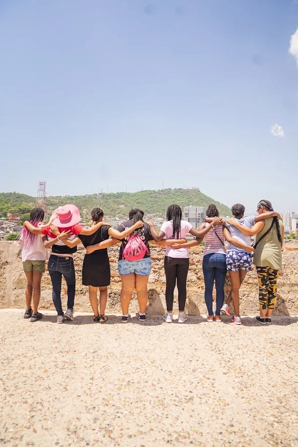 Ten women standing outdoors with arms around each other's shoulders, facing a cityscape and hill in the distance under a blue sky.