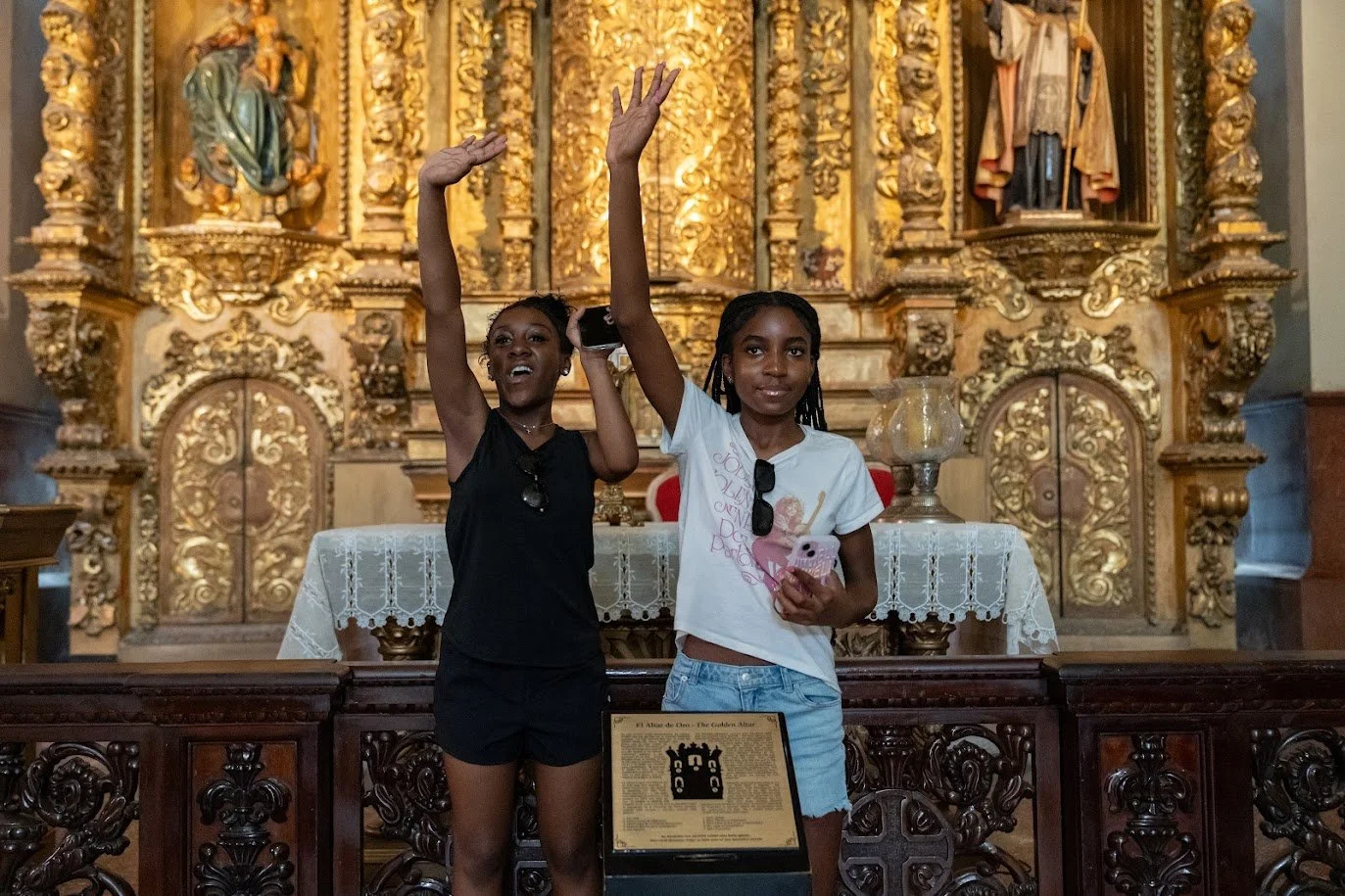 Two young girls standing inside a church with an ornate gold altar in the background. One girl in a black outfit is raising her right hand and smiling, the other in a white t-shirt and denim shorts is slightly smiling and holding a phone and a pink item. They are behind a wooden railing with a plaque, and a lace-covered table is in front of them.