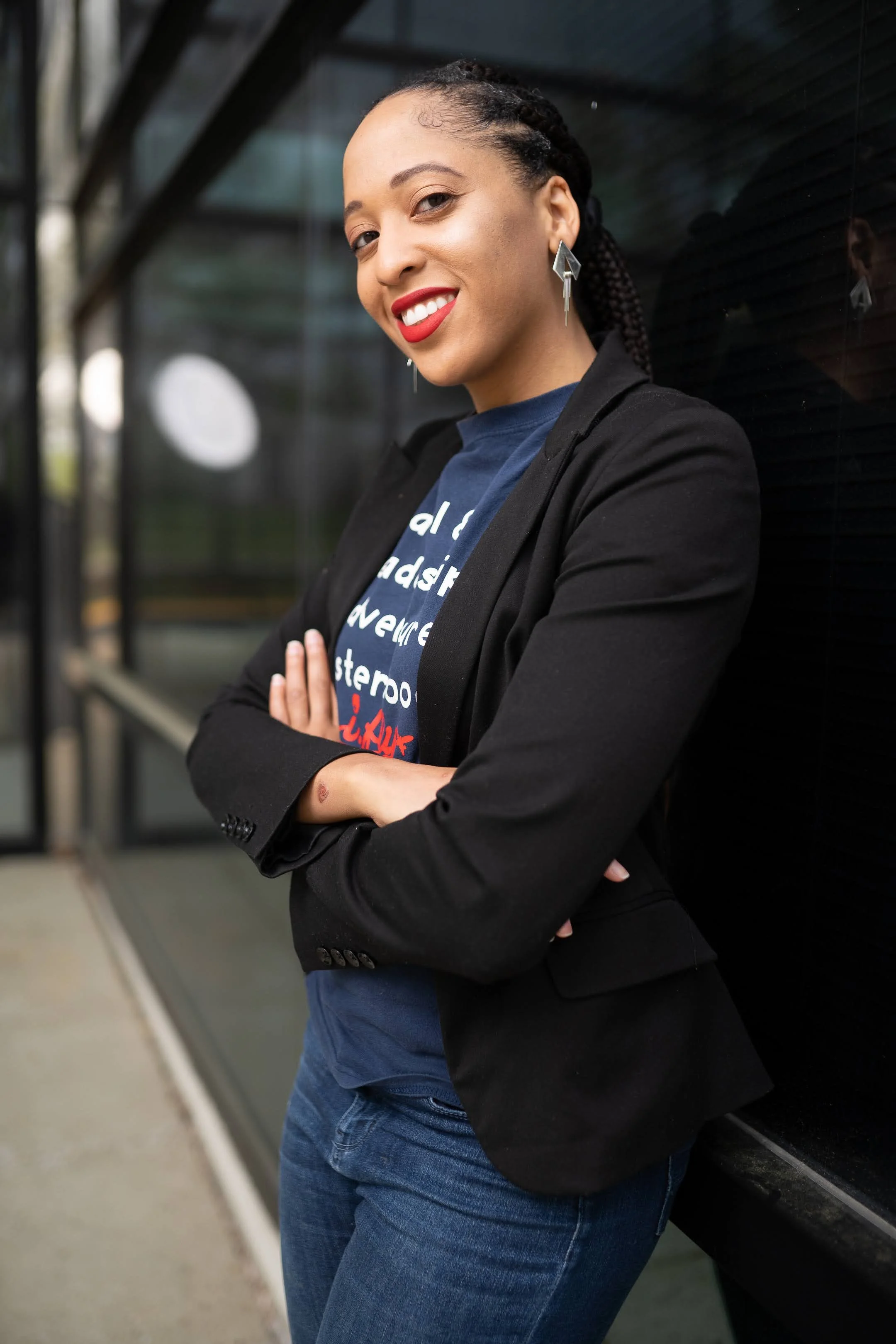 A confident woman with braided hair wearing a black blazer over a graphic t-shirt, standing with crossed arms outside against a dark wall, smiling with red lipstick.