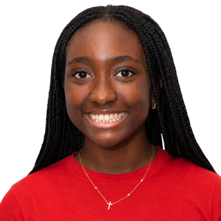A young woman with long braided hair smiling, wearing a red top and a gold necklace with a cross pendant.