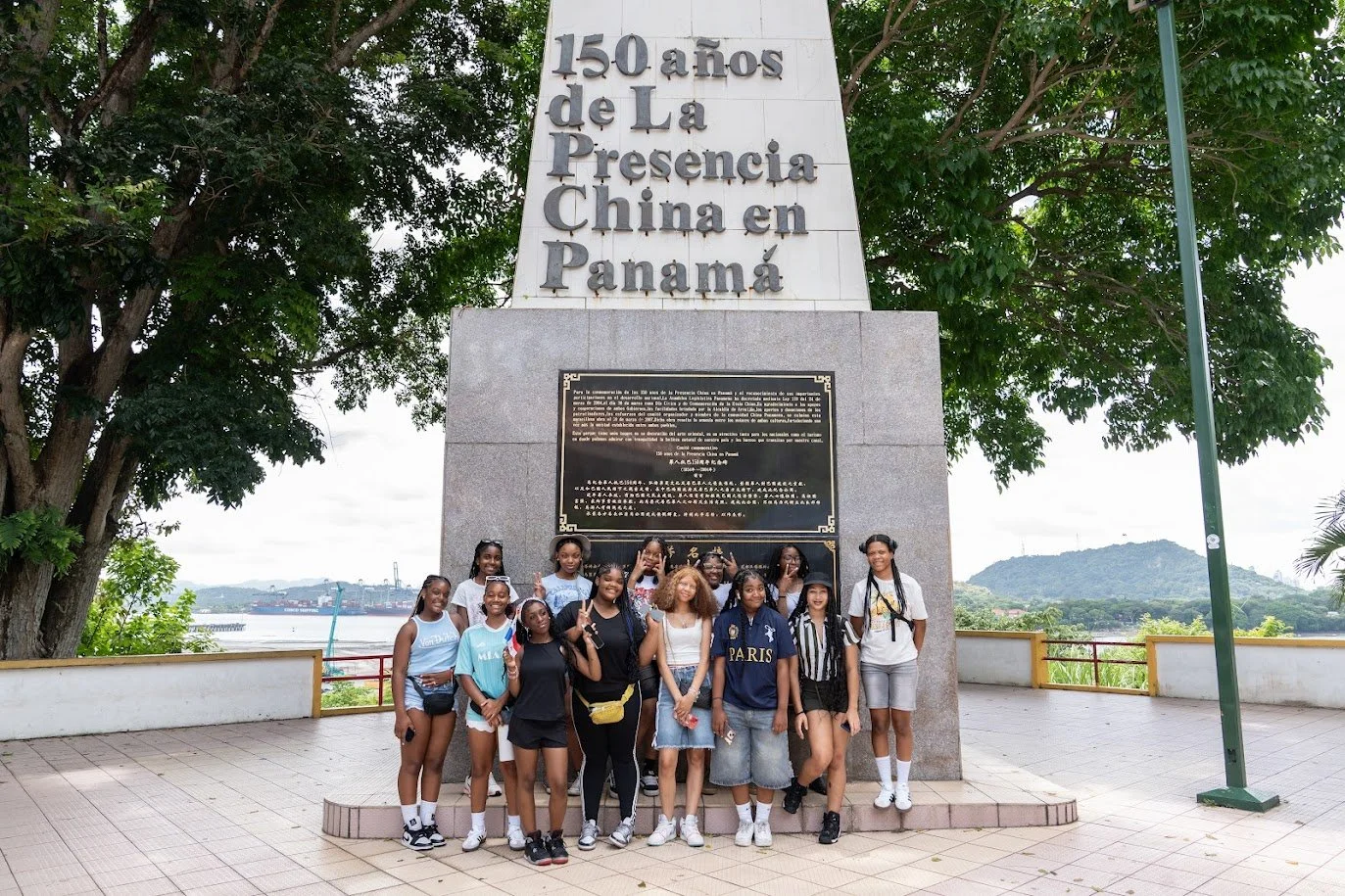 Group of children and teenagers standing in front of a monument with a plaque, celebrating the 150th anniversary of China's presence in Panama, with trees and mountains in the background.