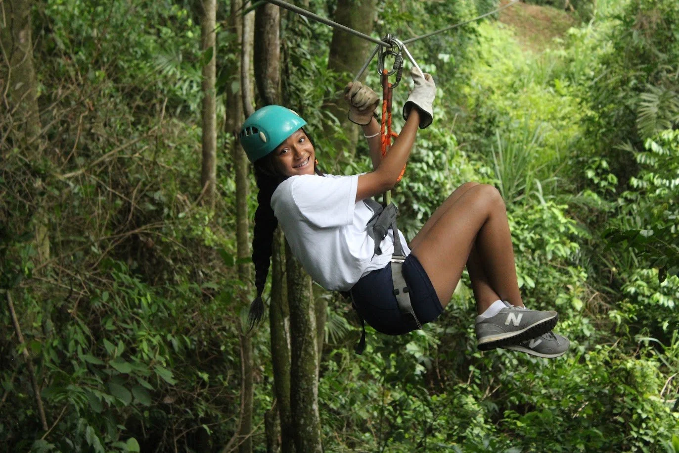 Young girl with a helmet and gloves on a zip line through a lush green forest, smiling at the camera.