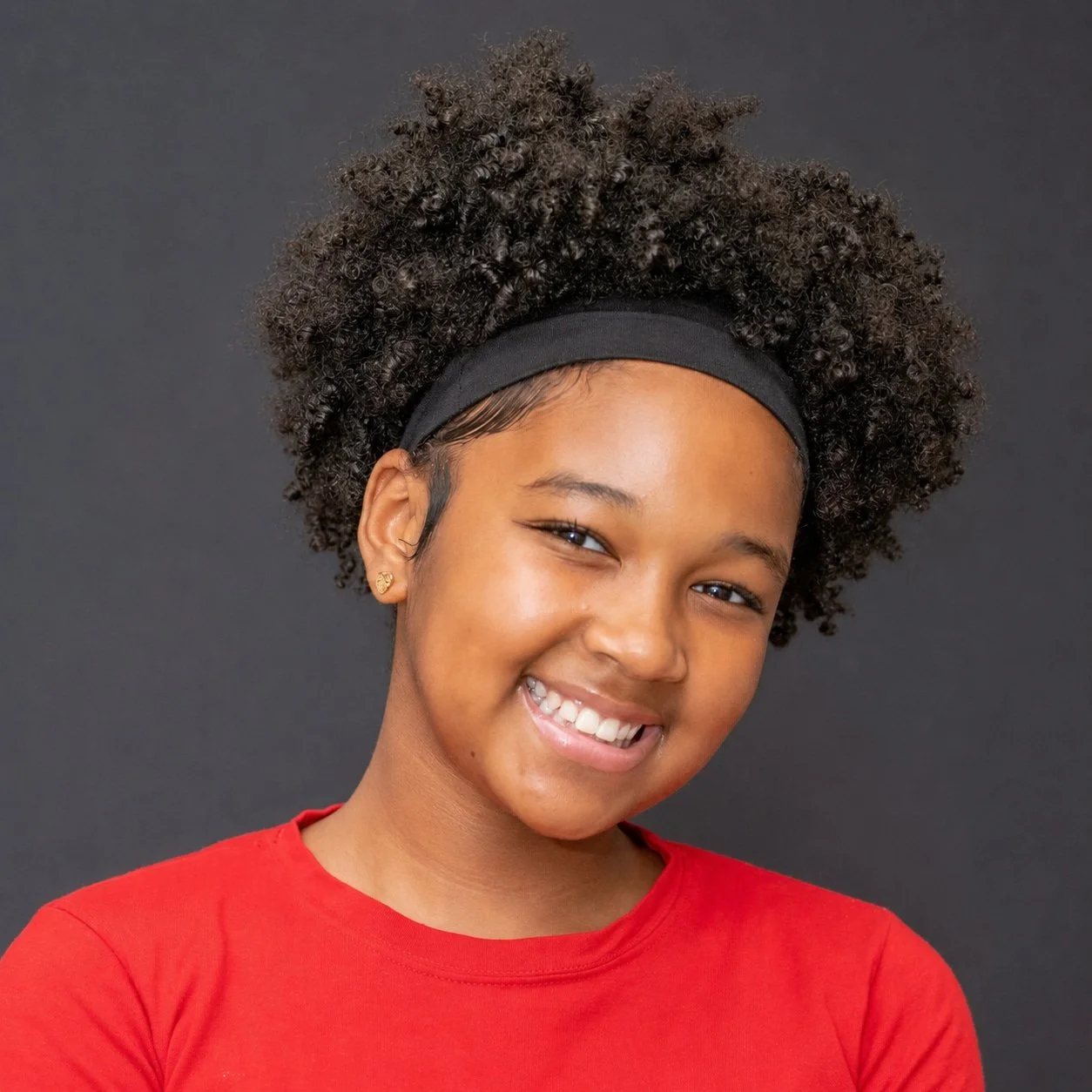 Smiling girl wearing red shirt and black headband against dark background.