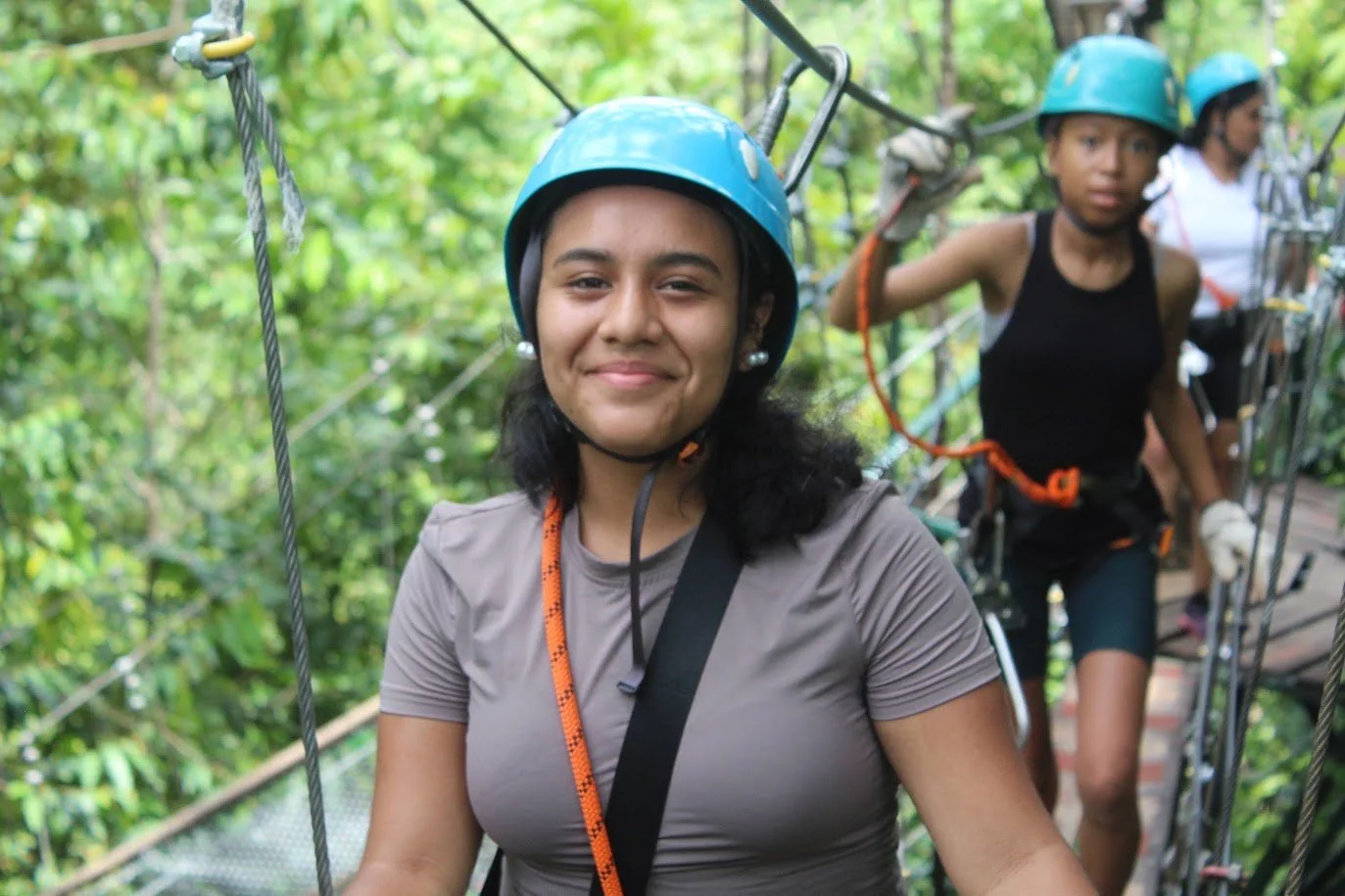 Group of people wearing helmets on a zipline in a lush green forest.