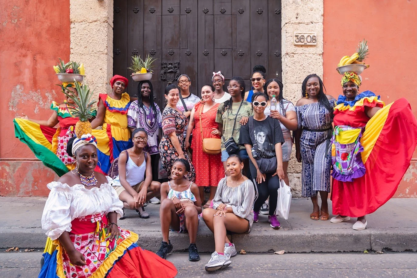 Group of women and girls with some dressed in vibrant traditional attire, some wearing accessories, standing and sitting in front of a large wooden door on a city street.