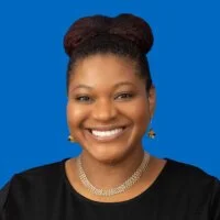 Portrait of a smiling woman with dark hair styled in an updo, wearing a black top, pearl necklace, and earrings, against a blue background.