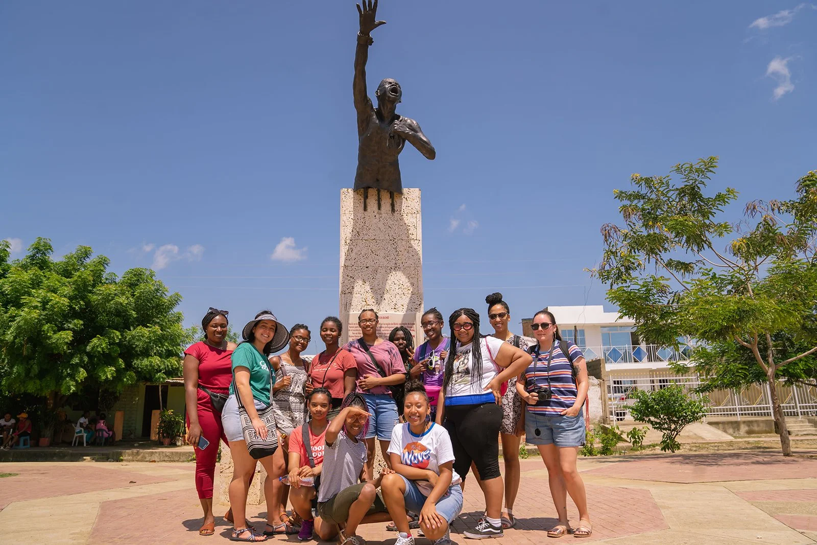 Group of diverse women and girls smiling and posing in front of a statue of a woman with raised arm, outdoors on a sunny day, with trees and buildings in the background.
