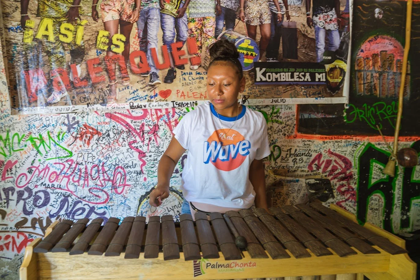 Young girl playing a wooden marimba in front of a colorful graffiti-covered wall with various handwritten messages and images, including a large poster with the phrase 'Ya Es Valer Que!'