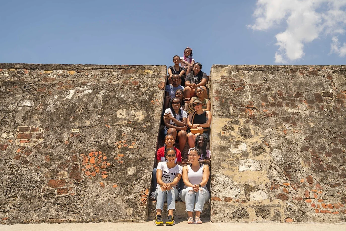 A group of people sitting on an ancient stone staircase built into a wall, outdoors under a blue sky with clouds.
