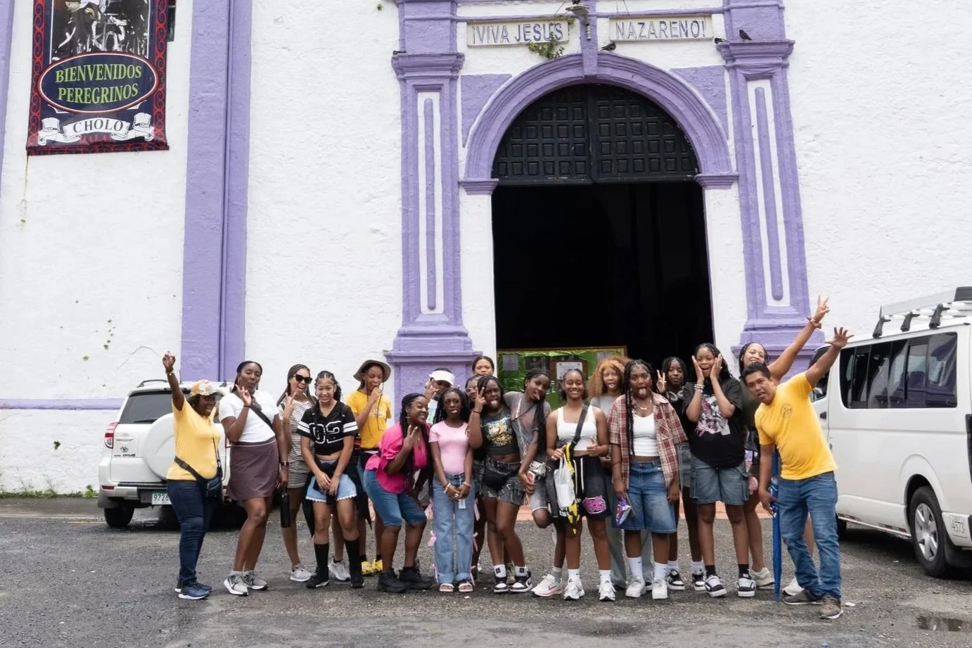 A group of young people standing in front of a white and purple church, posing for a photo and making peace signs. Two cars are parked behind them.