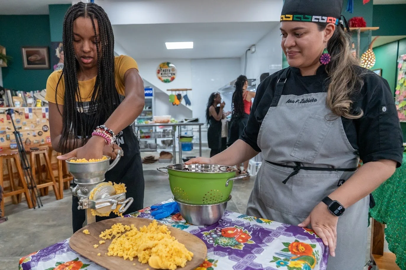 Two women preparing food in a kitchen or community space, with a table covered with a floral tablecloth, one woman wearing a yellow shirt and the other wearing a black shirt with a gray apron and a colorful headband.