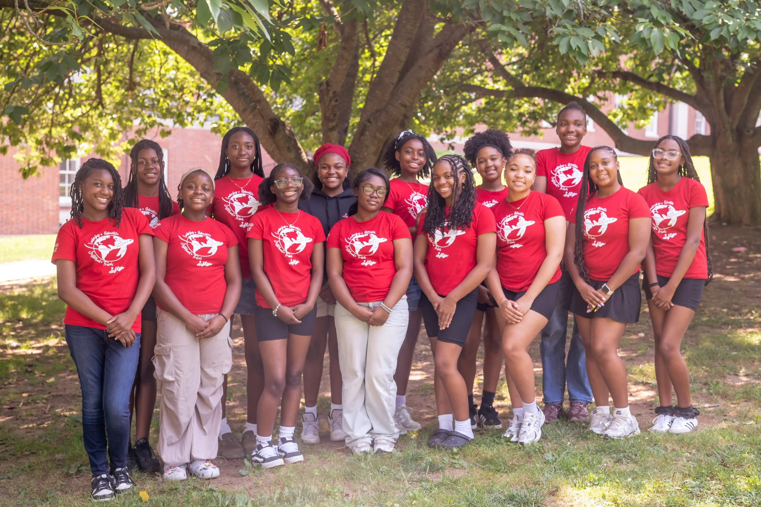 Group of young girls standing outdoors under a large tree, smiling at the camera, with some wearing red shirts featuring a white globe and plane logo.