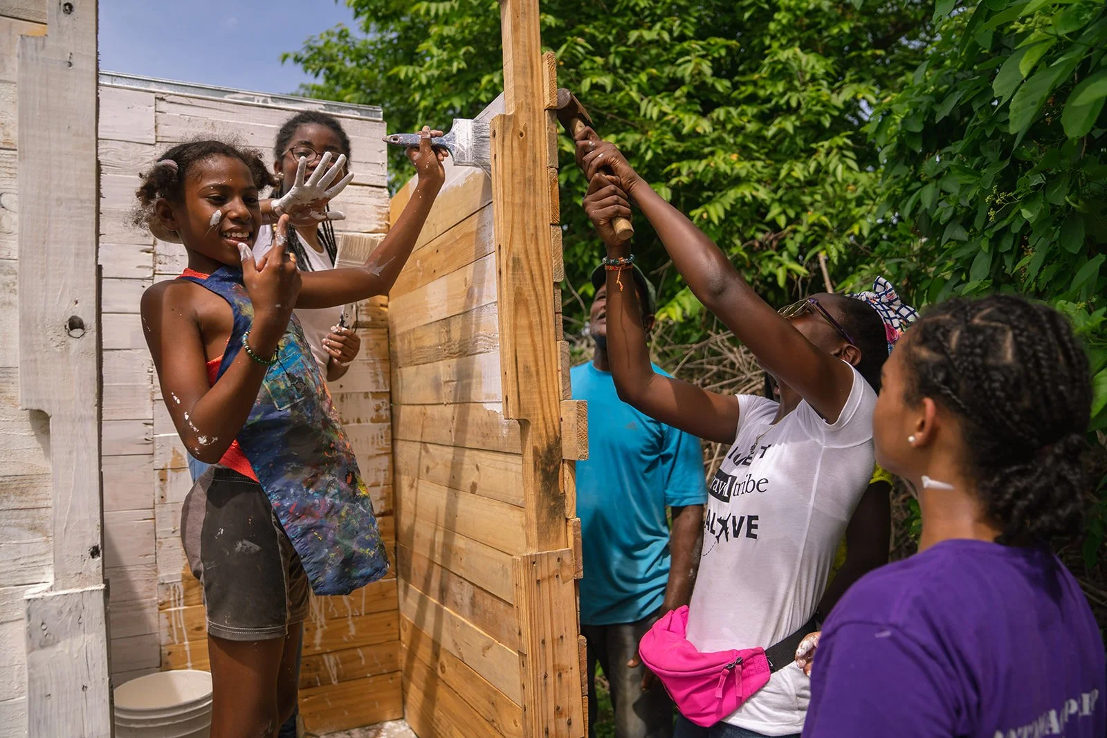 Group of women and girls building a wooden outdoor shower, with some applying paint and others working with tools.