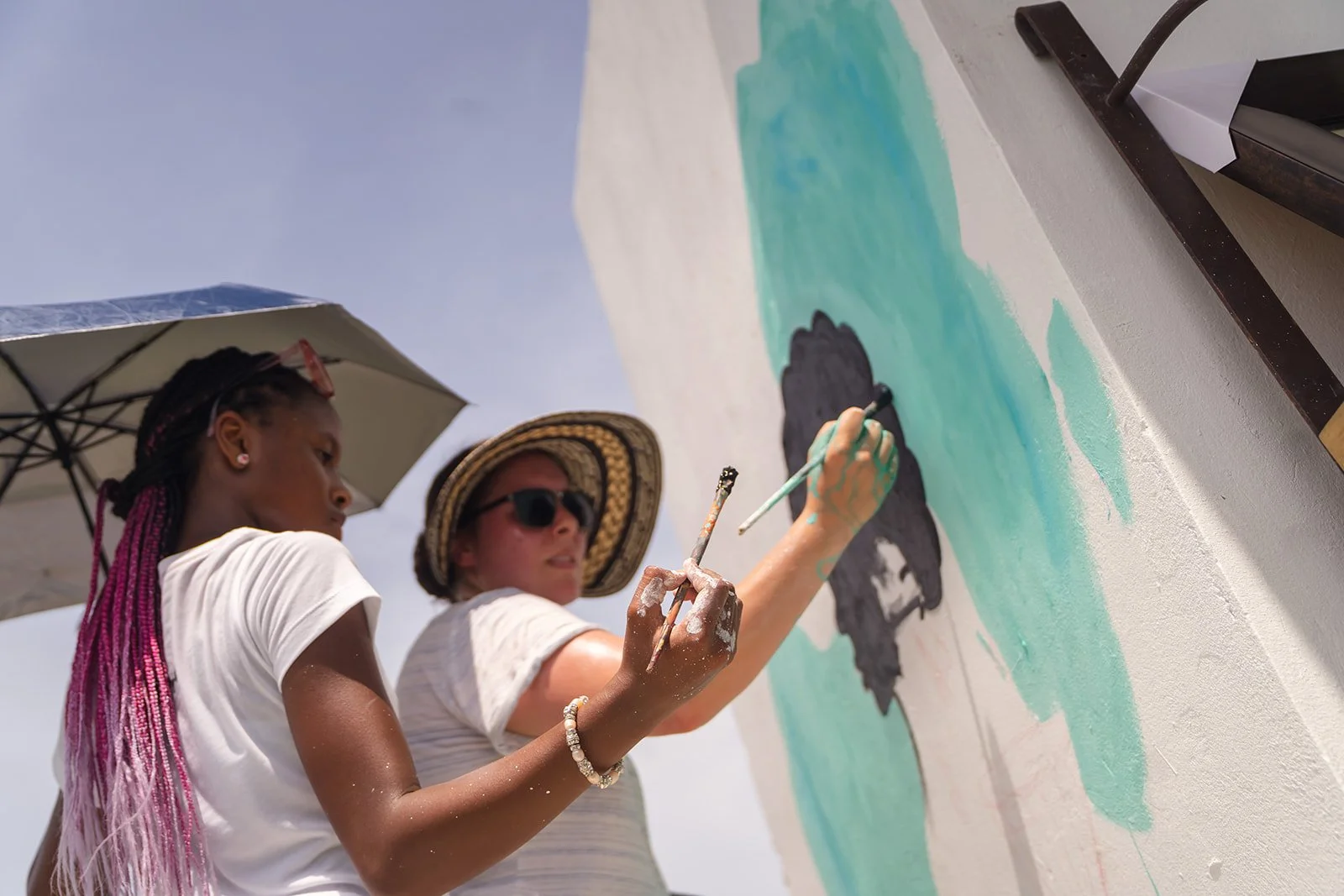 Two women painting a mural outdoors on a sunny day, one holding an umbrella for shade, both wearing hats and casual clothes.