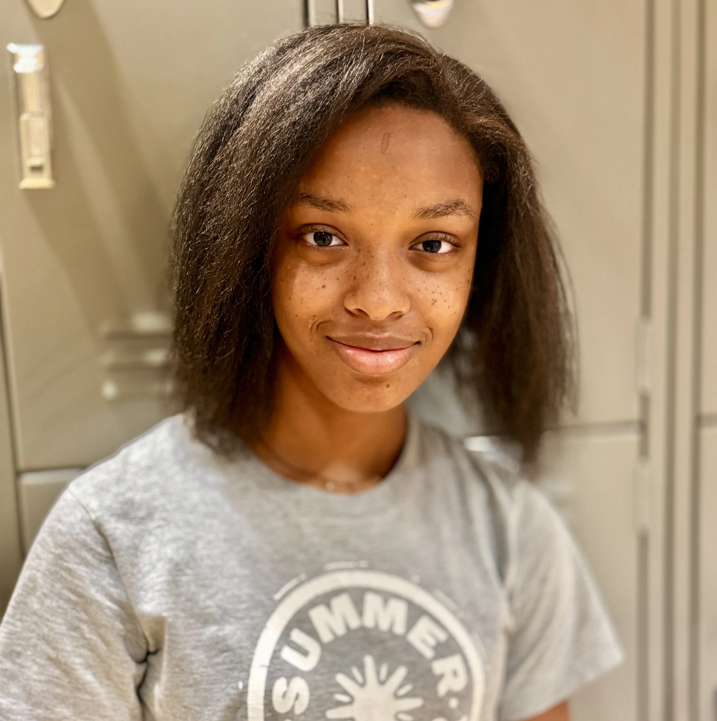 Close-up of a young woman with natural hair and brown skin, smiling at the camera, standing in front of a locker.