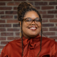 A woman with glasses and braided hair smiling in front of a brick wall.
