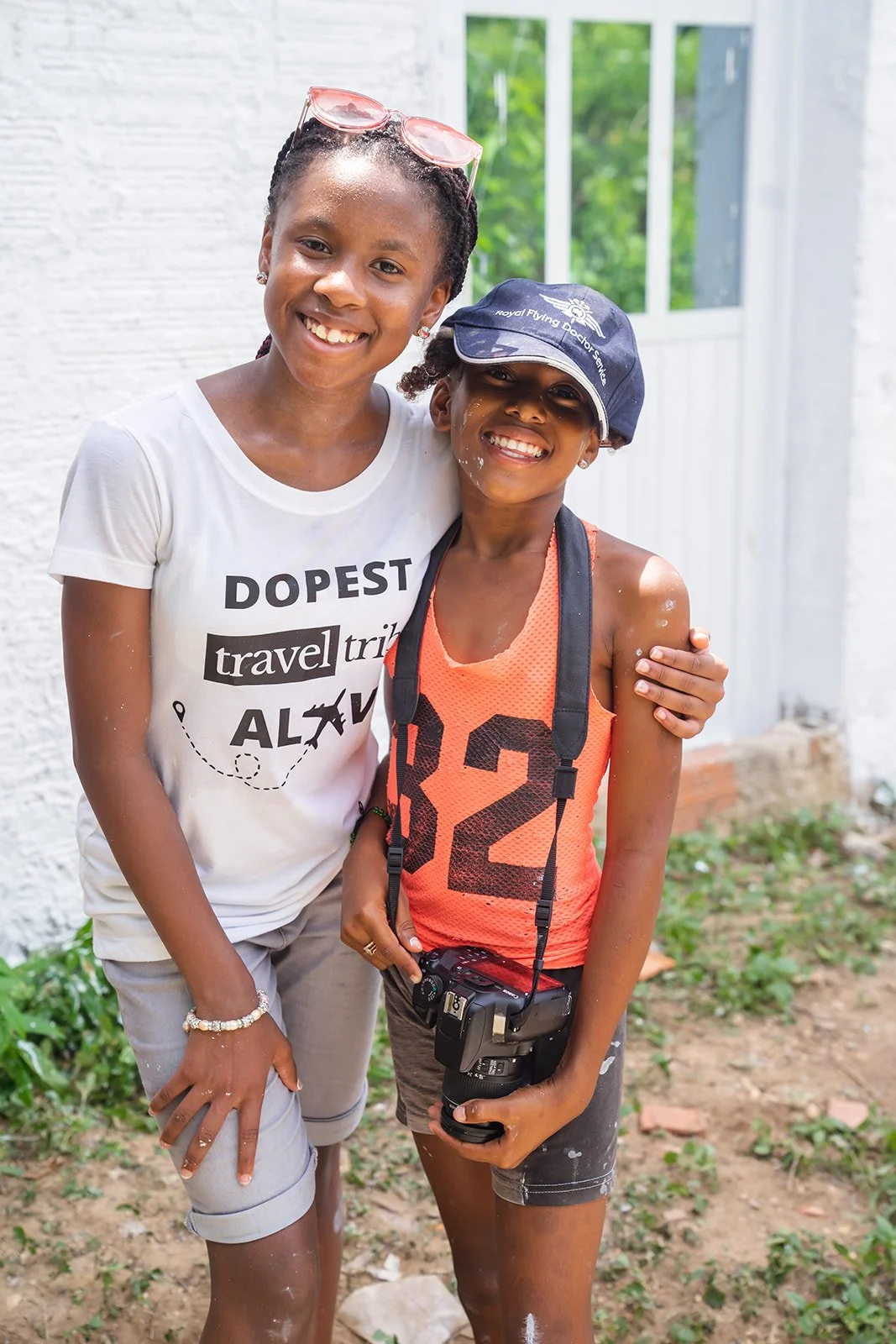 Two smiling women standing outside, one with her arm around the other. The woman on the left is wearing a white t-shirt with travel-related text and glasses on her head. The woman on the right is wearing an orange sports tank top with the number 32, a navy baseball cap, and a camera hanging around her neck. They are outdoors near a white brick building, with greenery in the background.