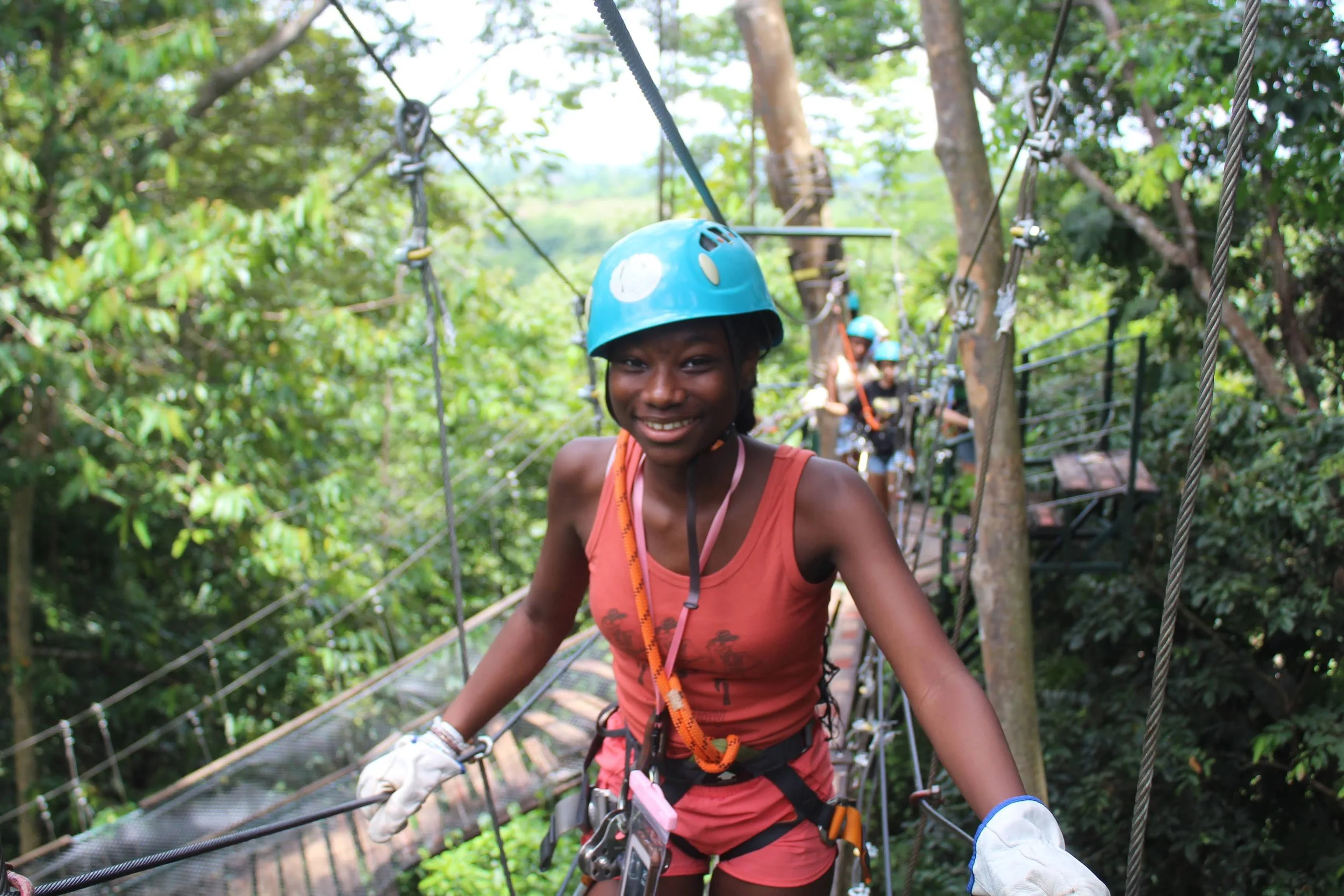 Woman smiling while walking on a suspension bridge in a forest, wearing a blue safety helmet and harness, with other people behind her also wearing helmets, participating in outdoor adventure activity.