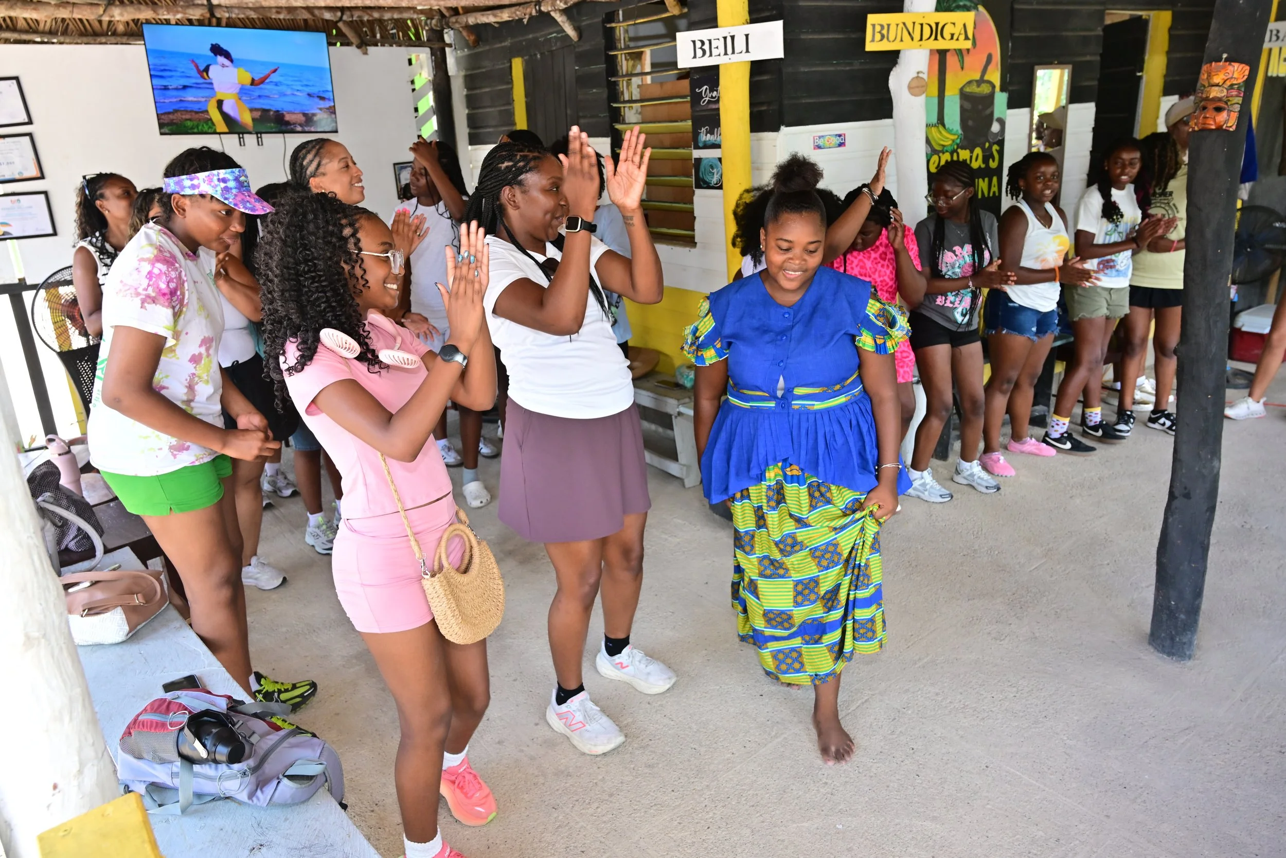 Group of women and girls standing in a line inside a decorated room, some with their hands raised and others clapping, while a woman in traditional African attire walks in front of them. The room has signs, artwork, and a television on the wall.