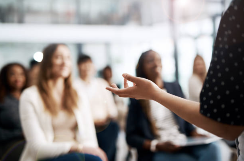 A woman speaking to an audience of diverse women in a modern, well-lit room.