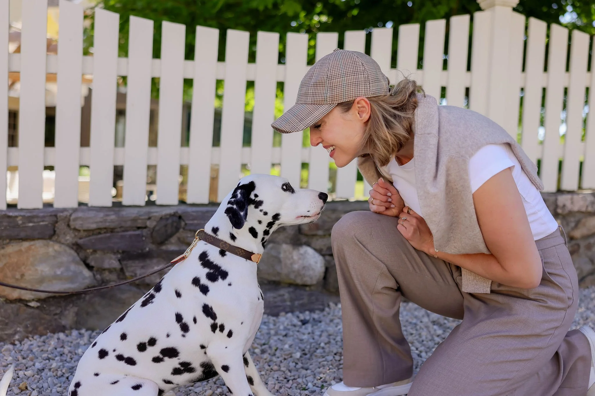 A woman with gray hair, wearing a plaid baseball cap and beige vest, crouches down smiling at a Dalmatian dog with black spots, in front of a white picket fence.
