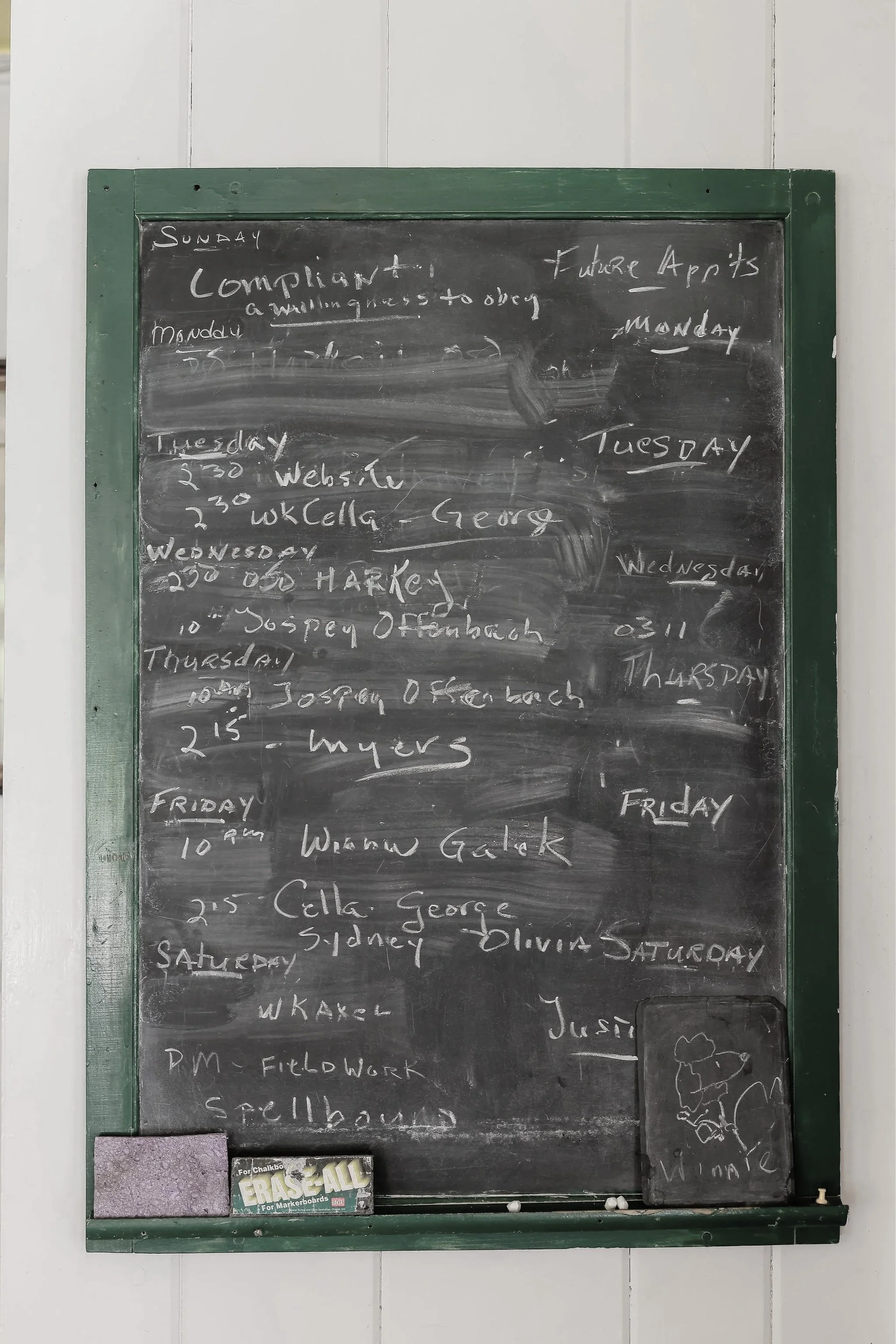 A black chalkboard with a green frame displaying weekly schedule written in white chalk, with a small pack of erasers at the bottom.