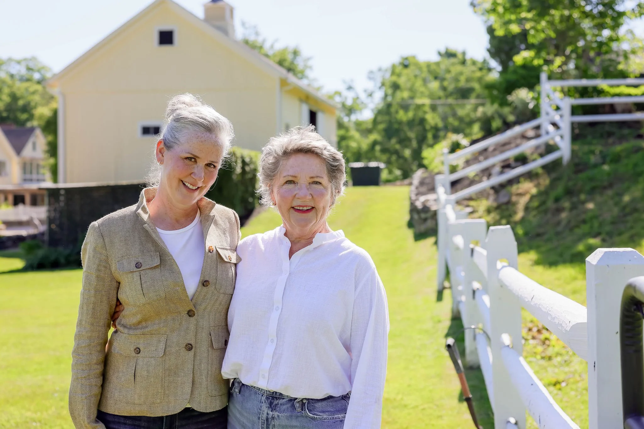 Two smiling women, possibly friends or relatives, standing outdoors in front of a white fence, with a yellow house and green trees in the background.