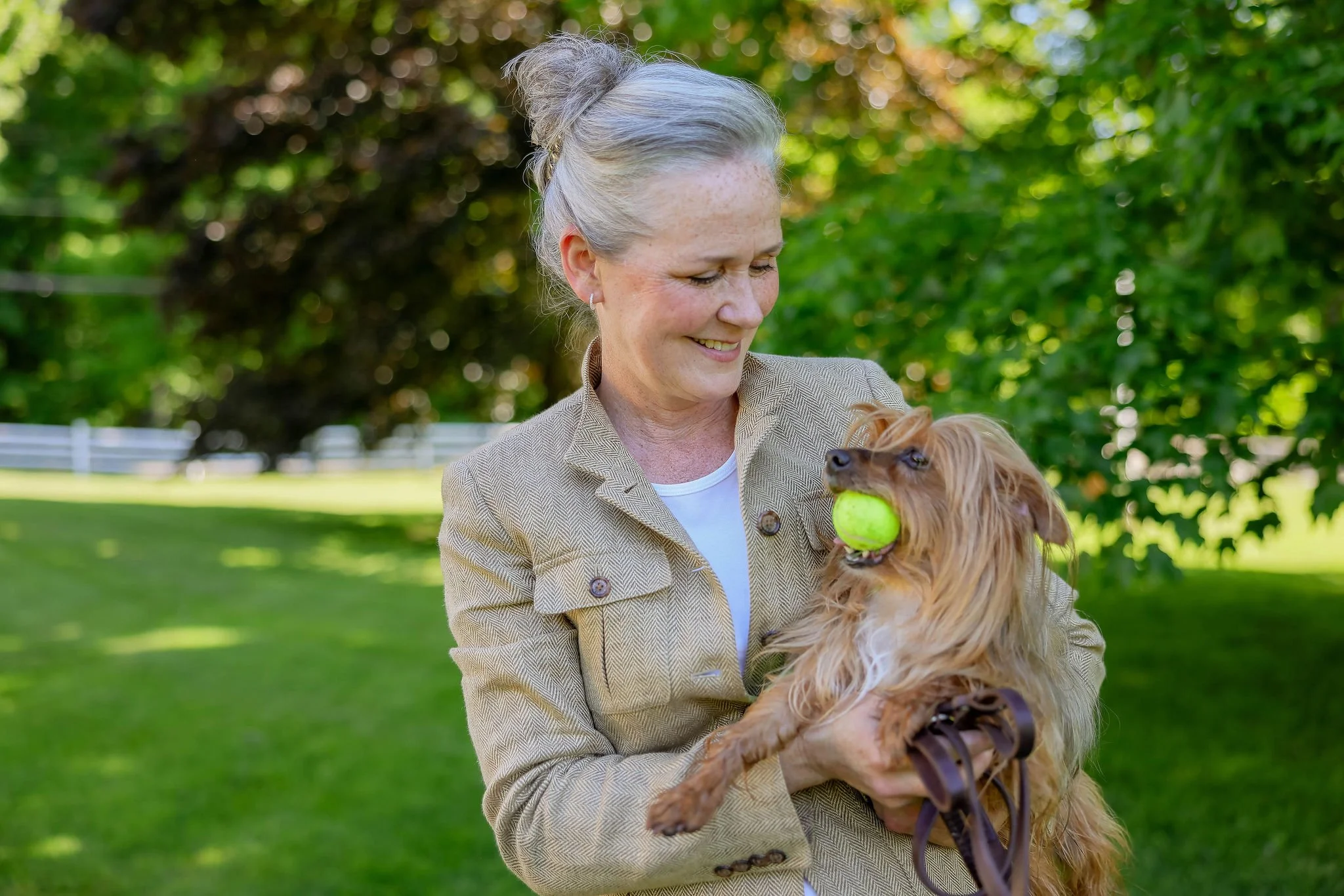 An elderly woman with gray hair in a bun holding a small dog with a tennis ball in its mouth outdoors.