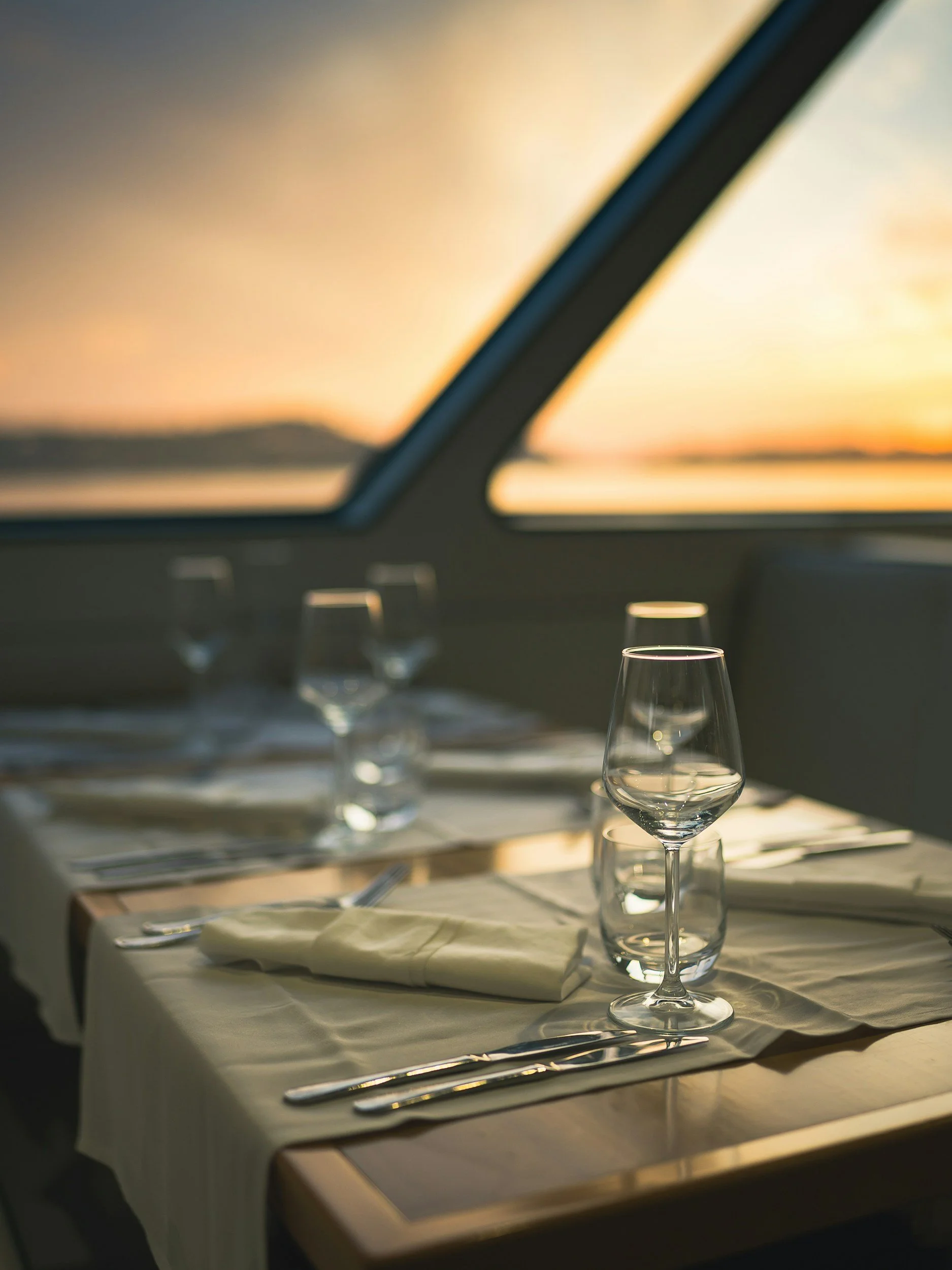 A table set for dining with stemmed glasses, cutlery, and folded napkins, illuminated by warm sunset light through a window with a blurred landscape view.