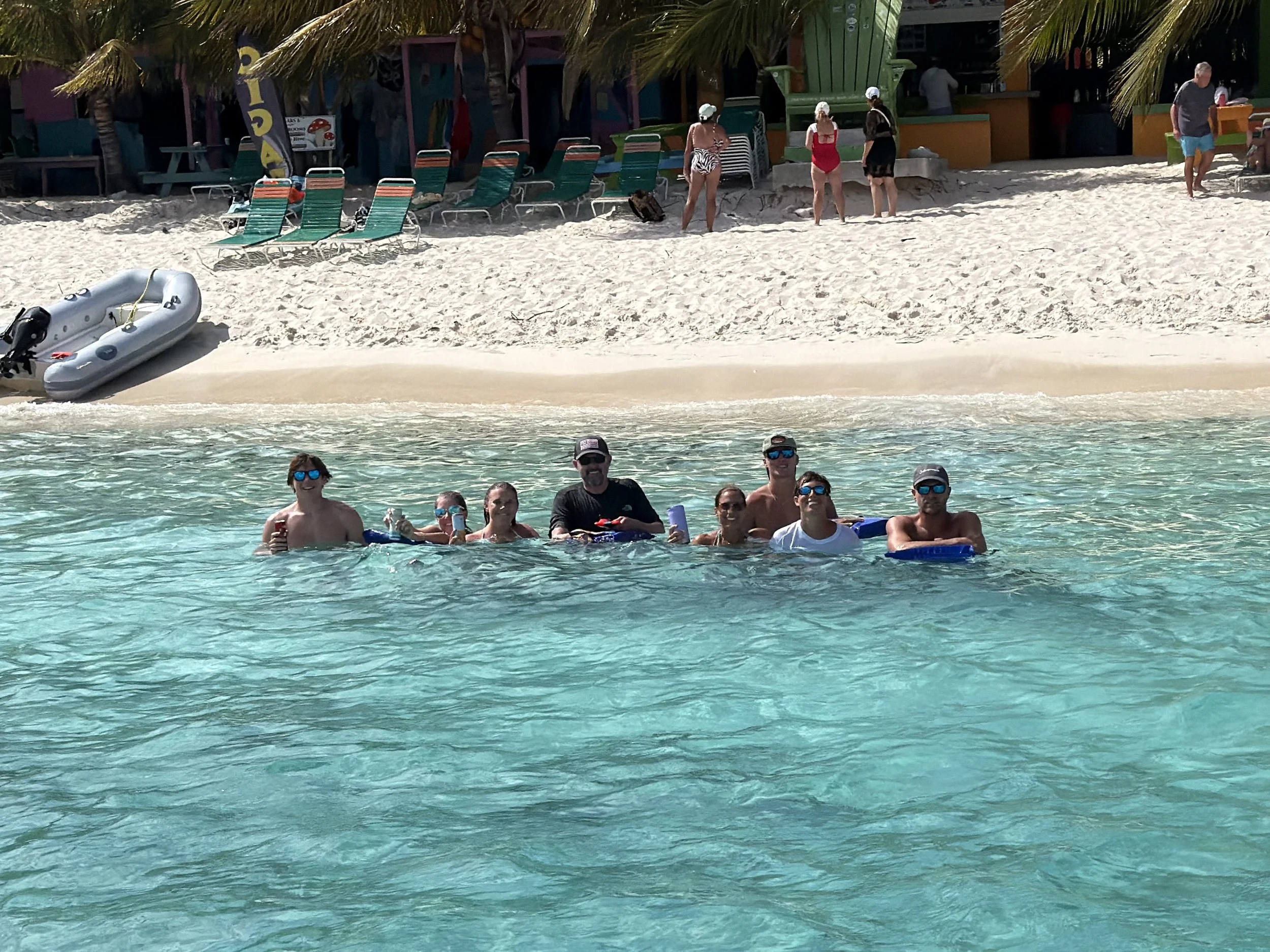 People enjoying time at the beach in the ocean, with some standing and others holding floatation devices. In the background, there are colorful chairs and palm trees near a beachside building.