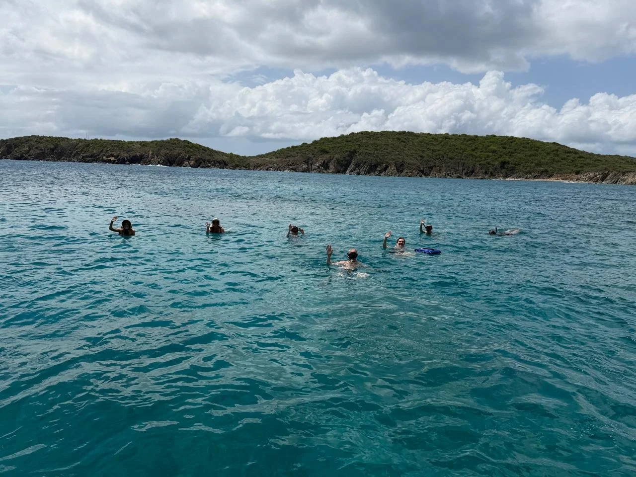 People swimming and waving in the ocean near a shoreline with green hills and cloudy sky.