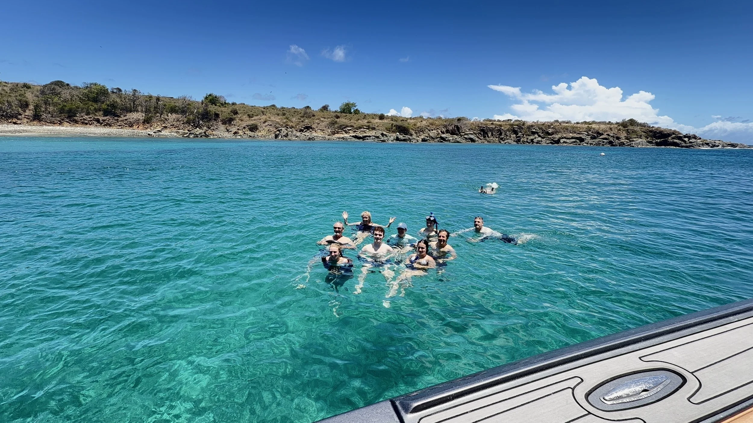 Group of people swimming and enjoying themselves in a clear blue body of water near a rocky shoreline, with a partly cloudy sky overhead.