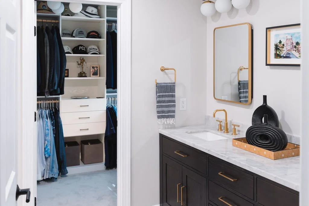 Bathroom with black vanity, white marble countertop, gold fixtures, wall mirror, gray towel, artwork, and decorative black oval vases. Adjacent walk-in closet with hats, trophies, framed photos, and clothing.