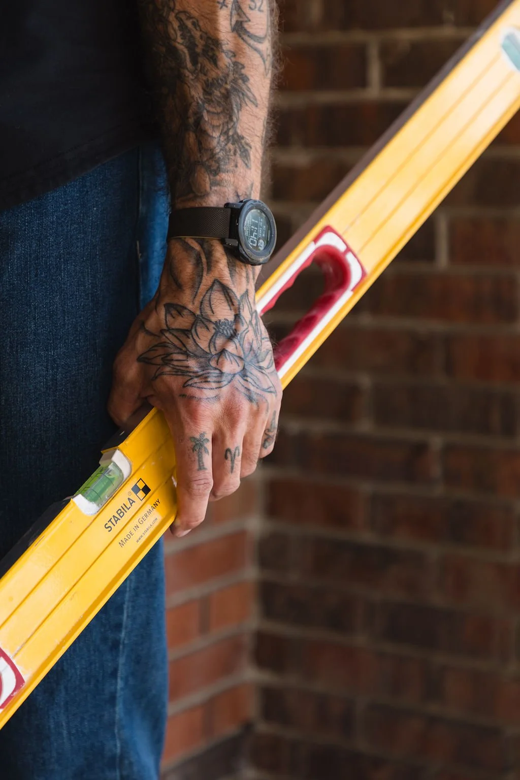 A tattooed person holding a yellow spirit level against a brick wall.