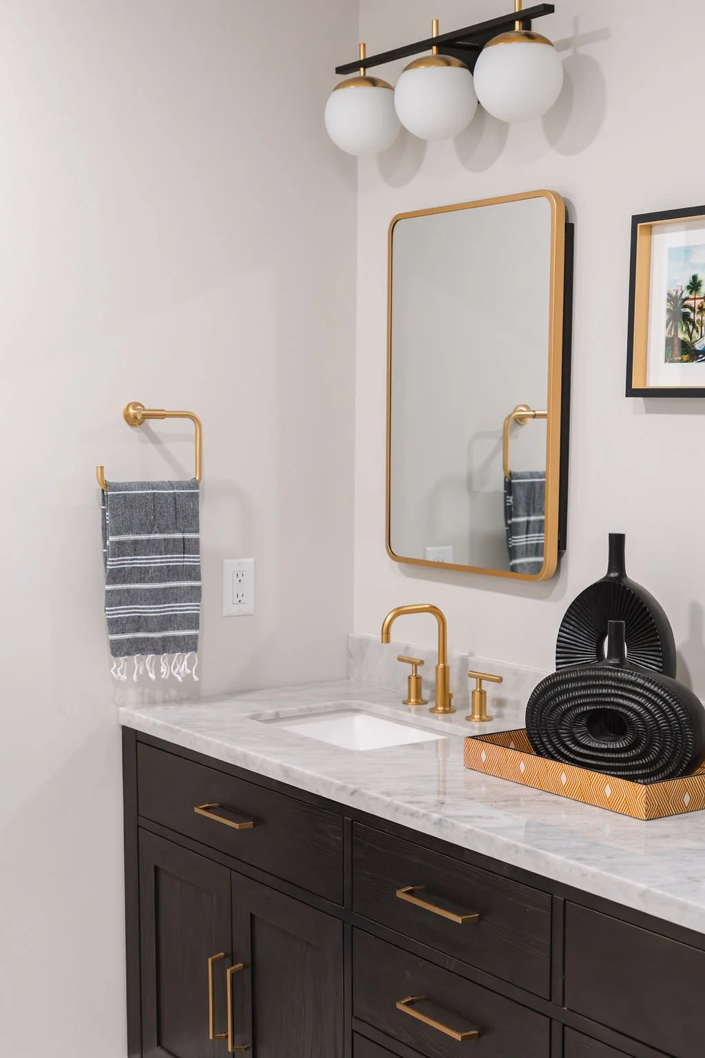 Modern bathroom vanity with dark wood cabinet, white marble countertop, brass fixtures, and decorative black vases on tray.