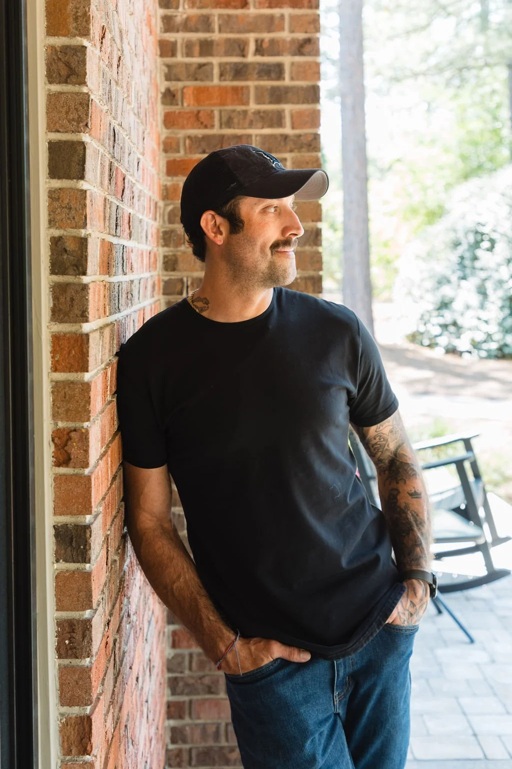 Jay Southers black t-shirt, and blue jeans leaning against a brick wall on an outdoor patio, with trees and outdoor furniture in the background.