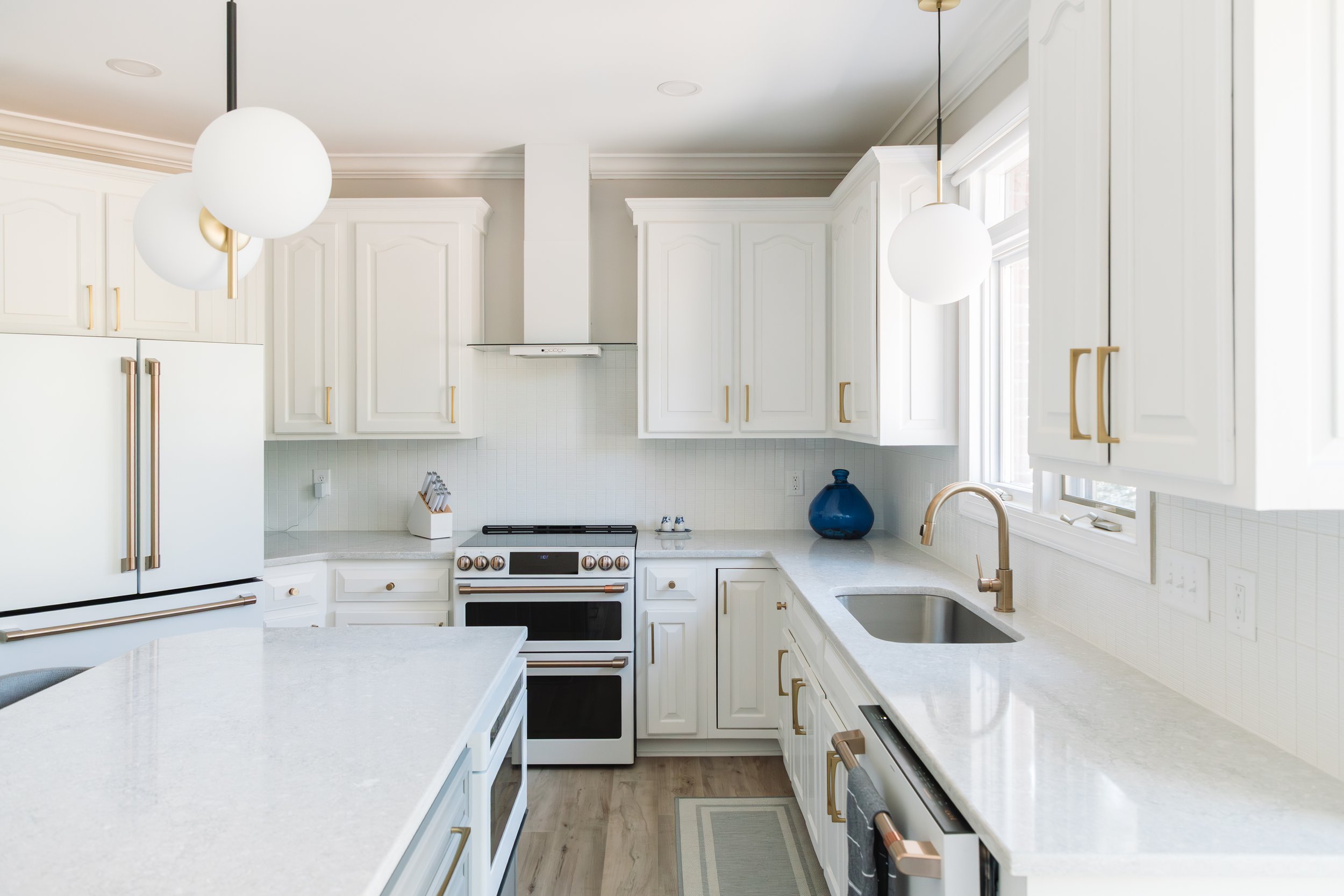 A modern white kitchen with white cabinets, gold hardware, a white stove, a stainless steel sink, large windows, and two white globe pendant lights with gold accents.