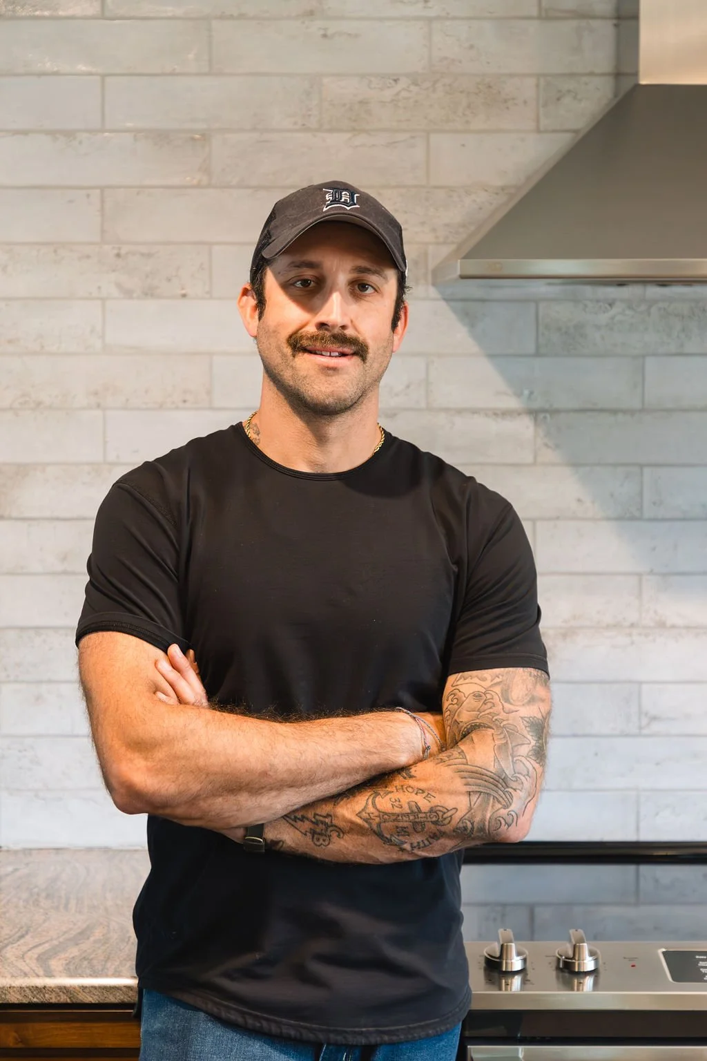 Owner and Operator, Jay Southerswearing a black cap and black t-shirt, standing with arms crossed in front of a renovated kitchen stovetop.