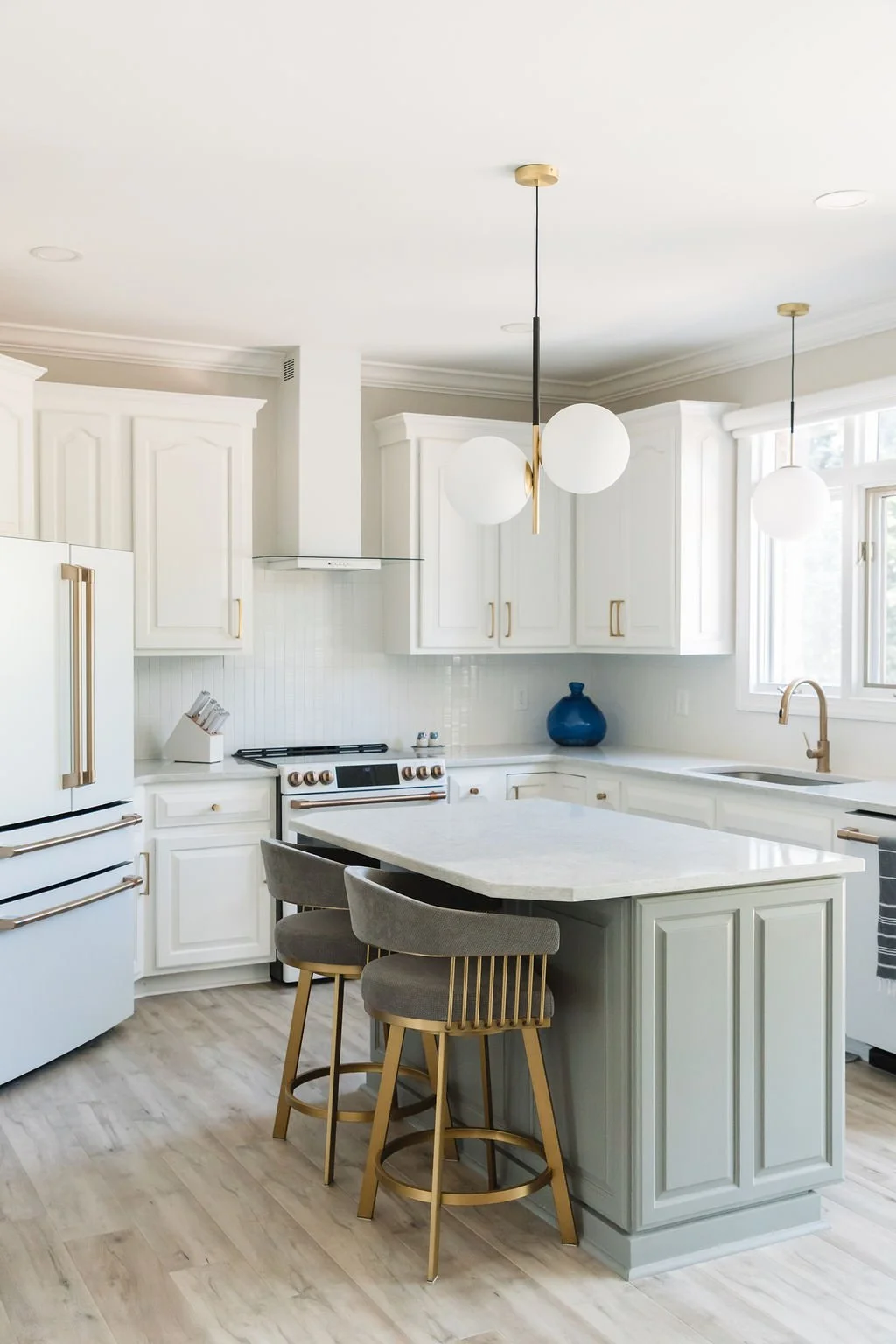 Modern white kitchen with gold accents, pendant lights, island with seating, white cabinets, and light wood flooring.
