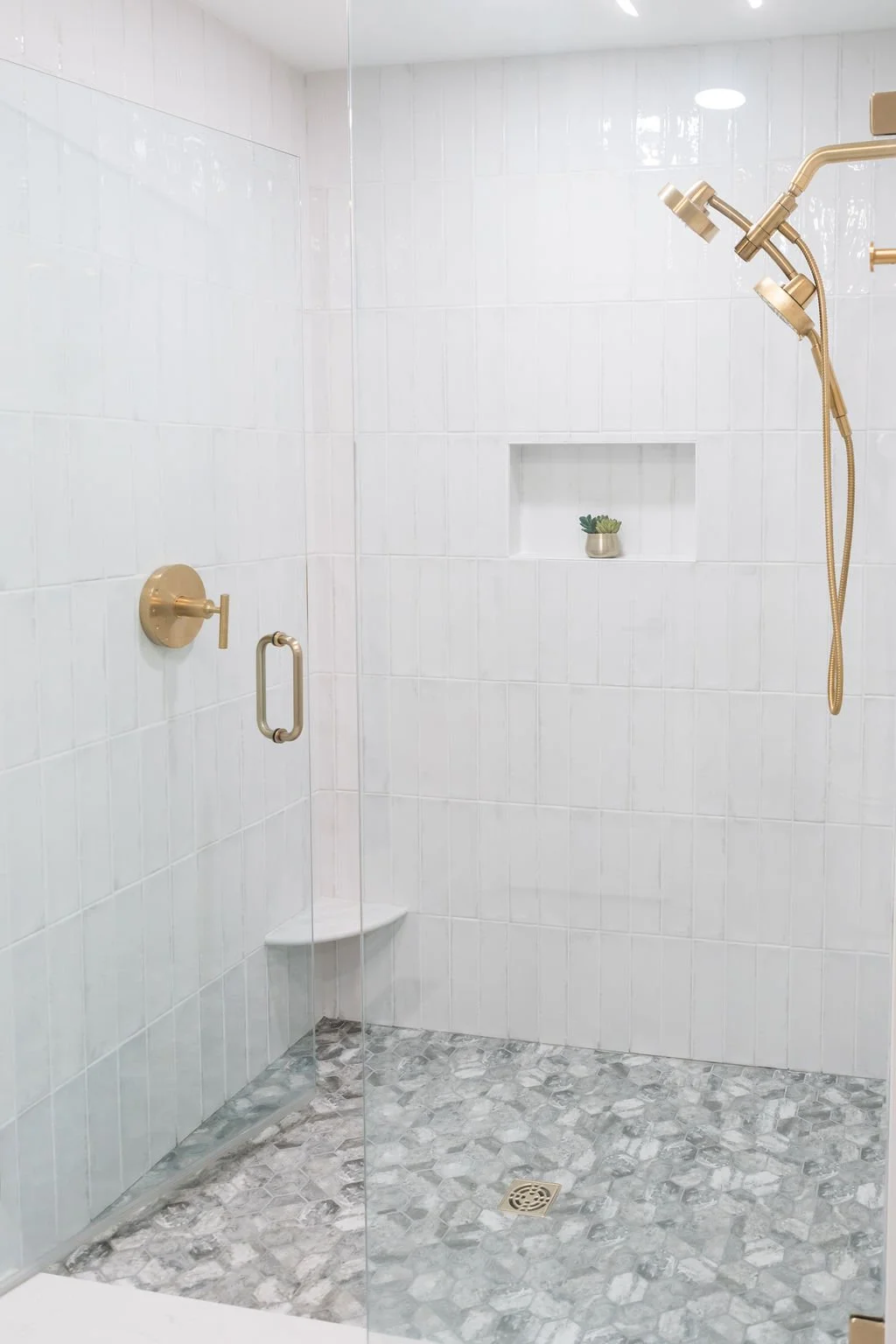 Empty walk-in shower with white tiled walls, gray patterned floor, gold fixtures, a built-in niche with a small plant, and a glass door.