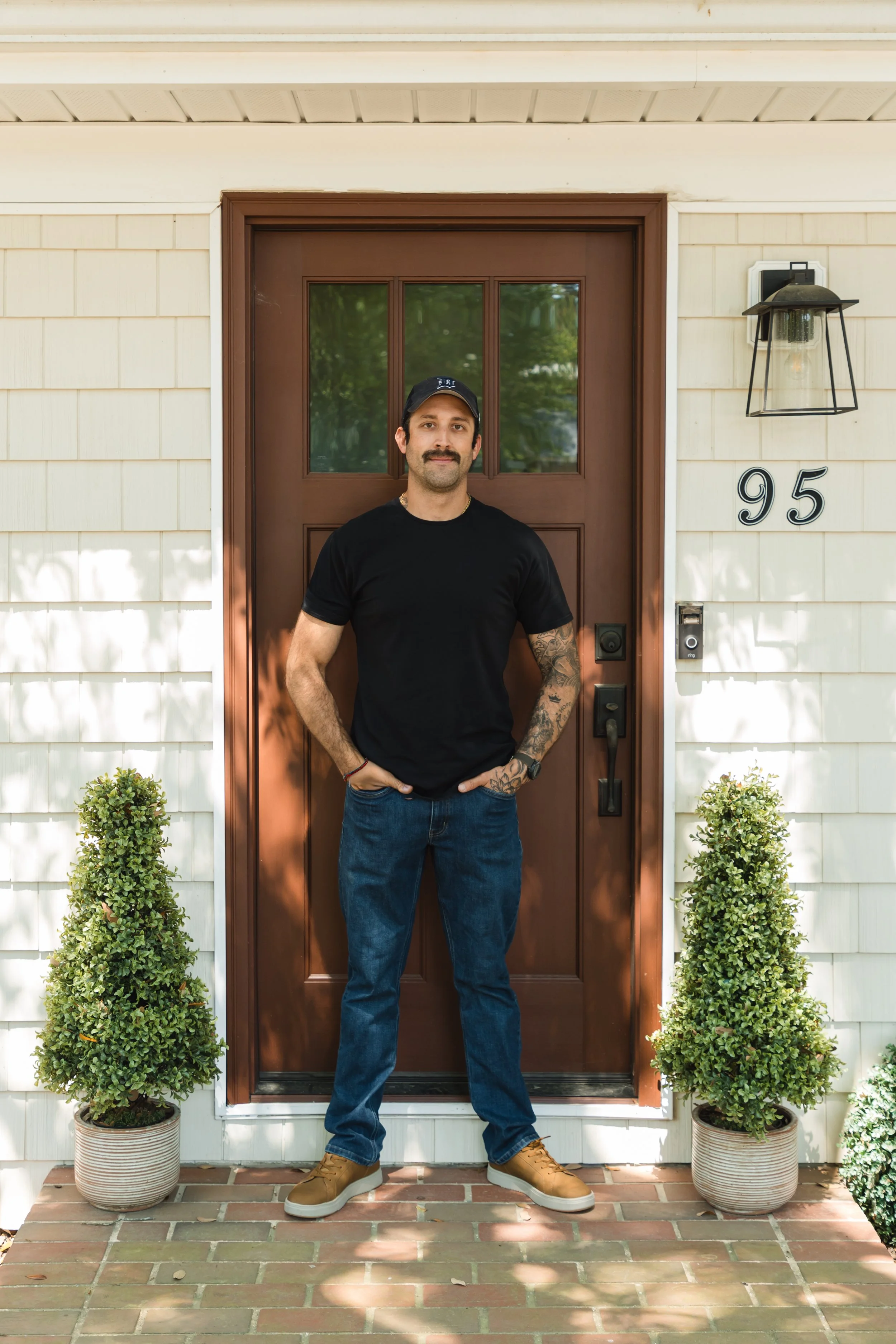 A man with a mustache, wearing a black t-shirt, blue jeans, tan shoes, a black cap, standing in front of a brown front door with the house number 95, flanked by two potted green shrubs.